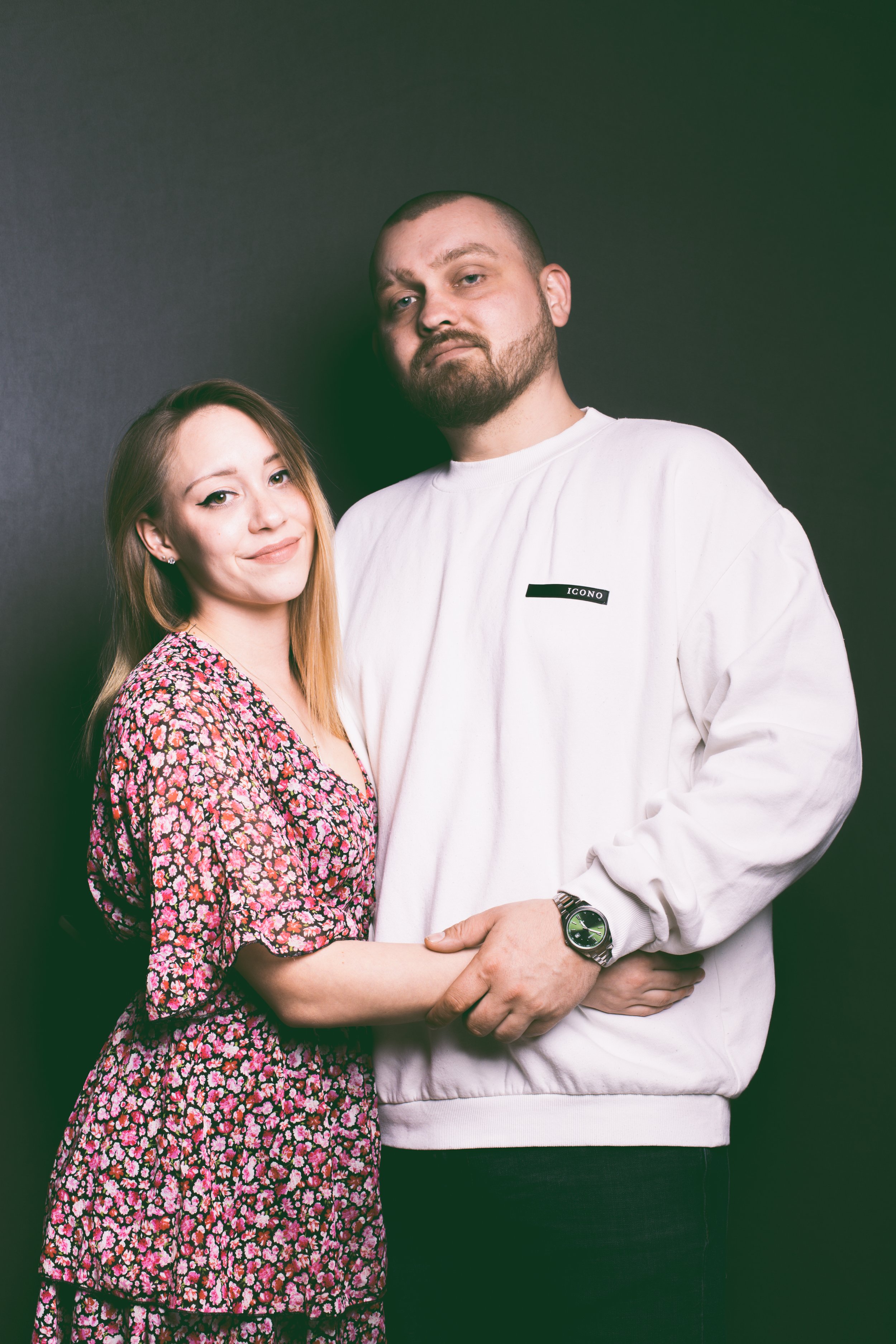 Ein junges Paar steht eng beieinander vor einem dunklen Hintergrund, beide schauen in die Kamera. Das Mädchen trägt ein blumiges Kleid, der Mann trägt ein weißes Sweatshirt mit einem kleinen schwarzen Etikett."ICONO" Es sind die Fotografen.