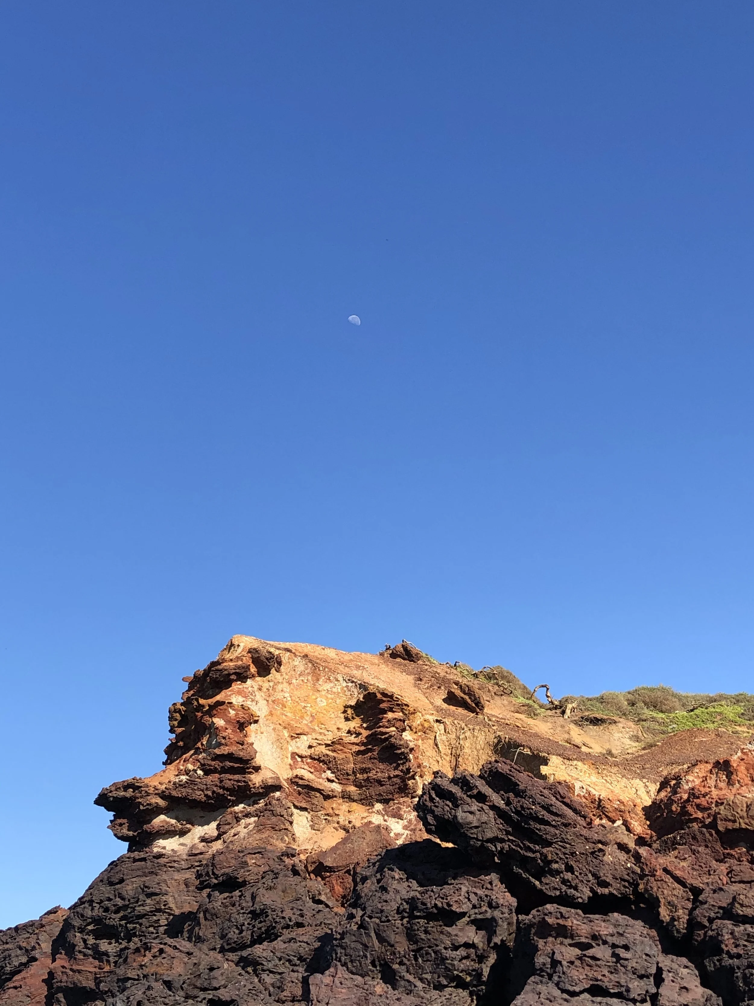 Close-up of a rugged, reddish-brown rock formation under a clear blue sky with a visible moon.