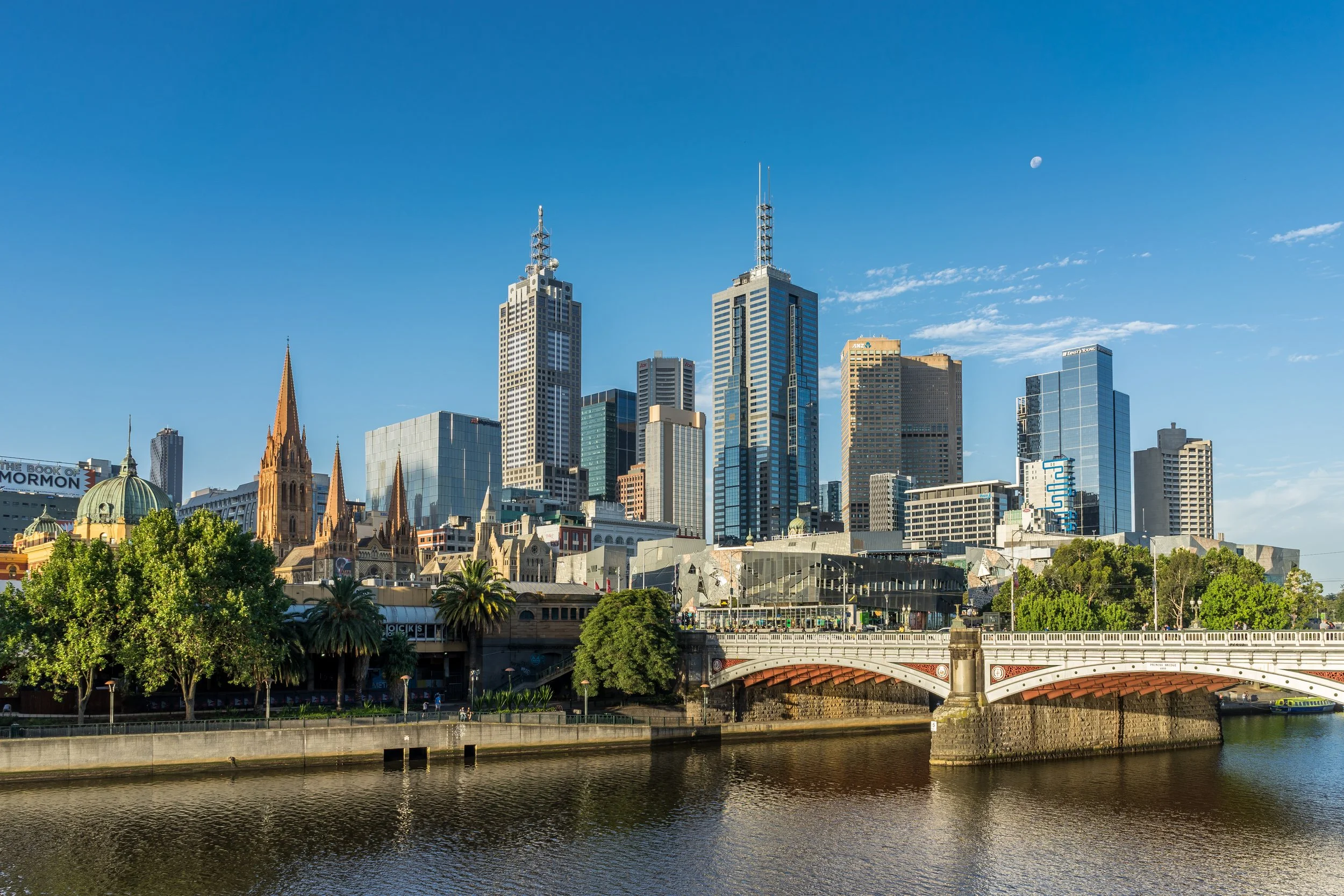 Skyline of downtown Melbourne, Australia, with modern skyscrapers, historic churches, arches of a bridge, and the river in the foreground on a clear day.