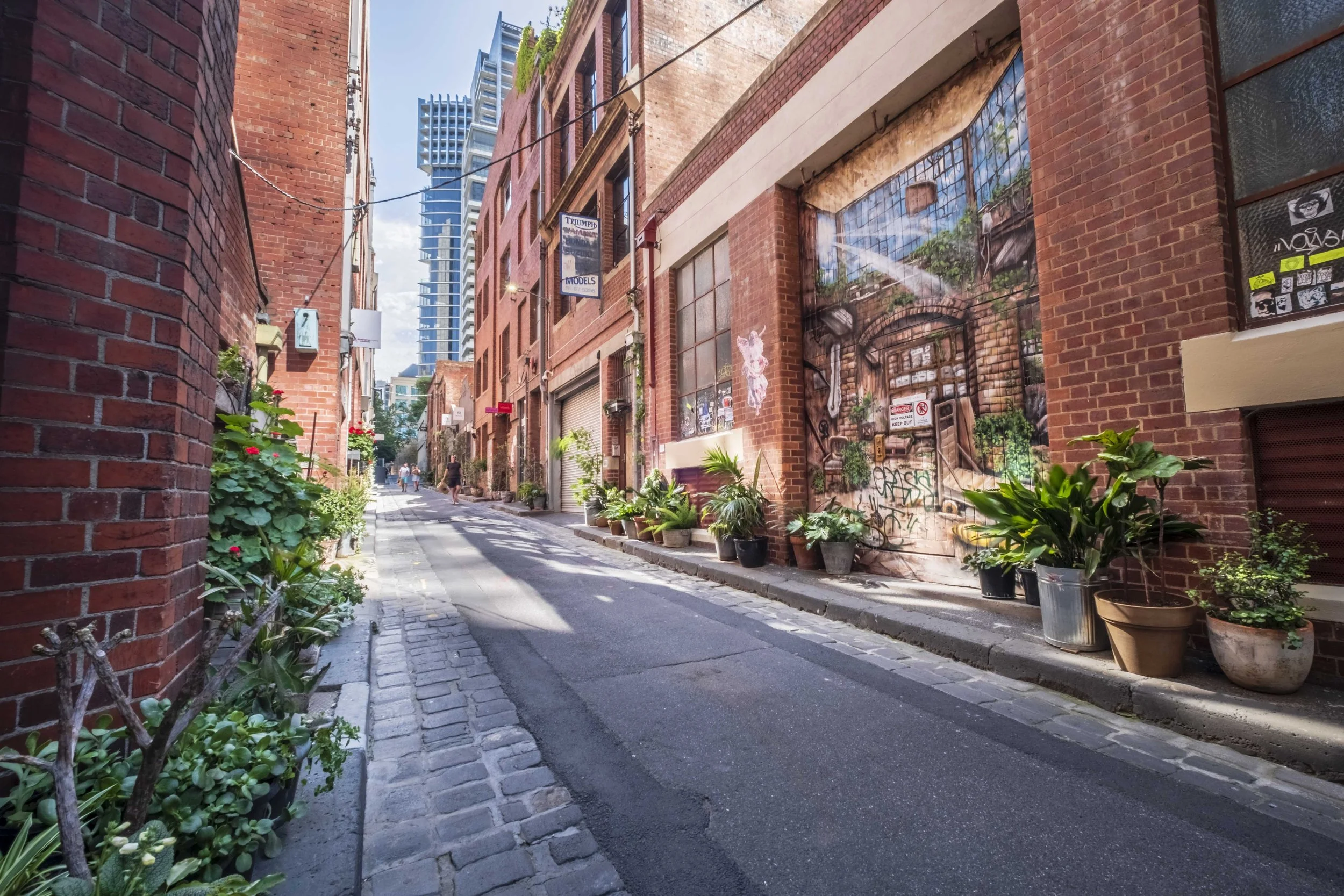 Urban alleyway in Melbourne CBD with red brick buildings, potted plants along the sidewalk, a mural on one building, and tall modern buildings in the background.