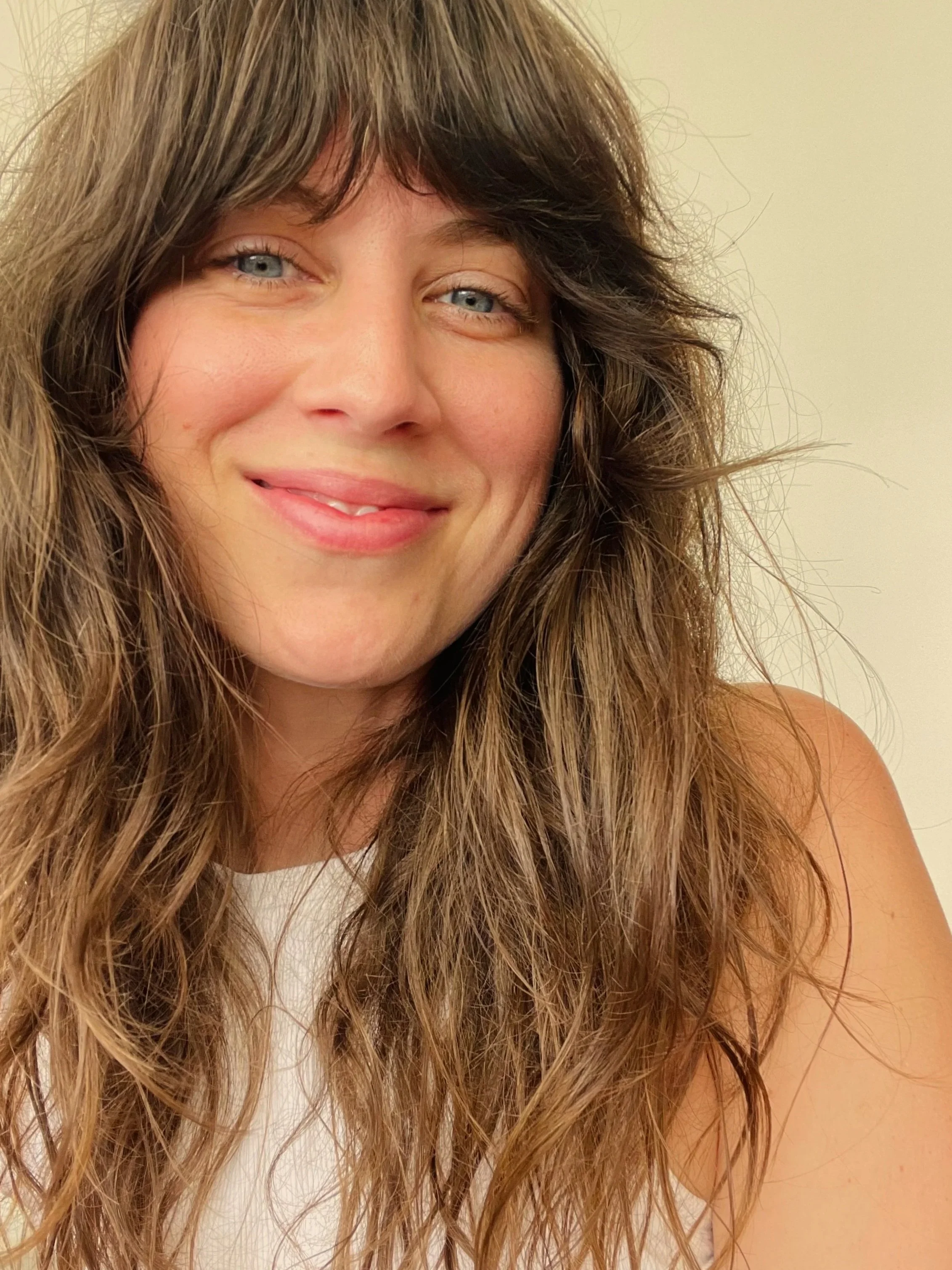 Close-up of a smiling woman with long, wavy brown hair and blue eyes, wearing a sleeveless white top.