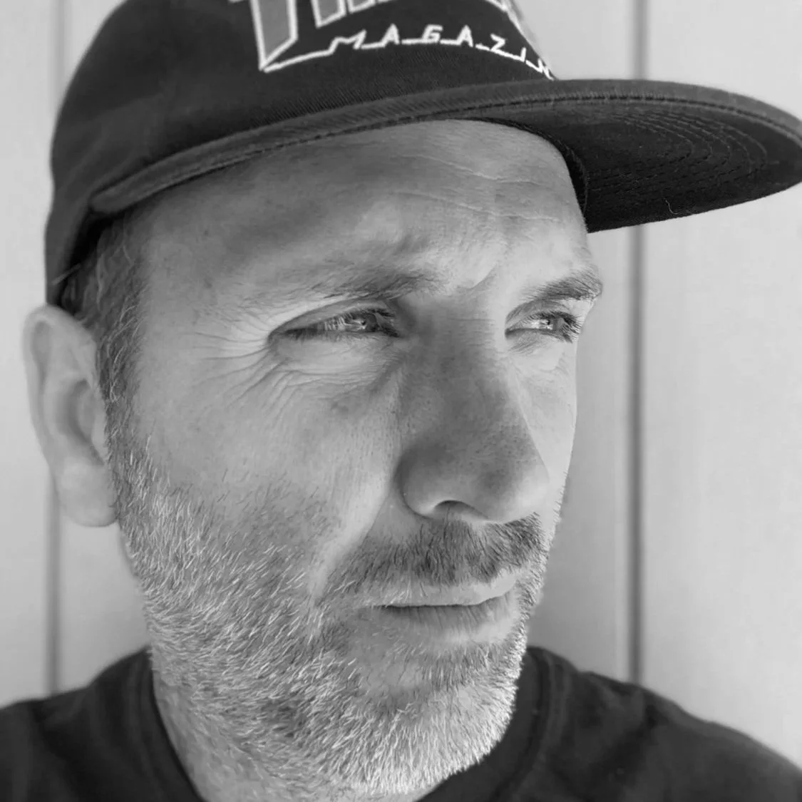 Black and white close-up photo of a middle-aged man wearing a baseball cap, looking slightly to the right. The man has a beard and mustache, with visible wrinkles and light-colored eyes.