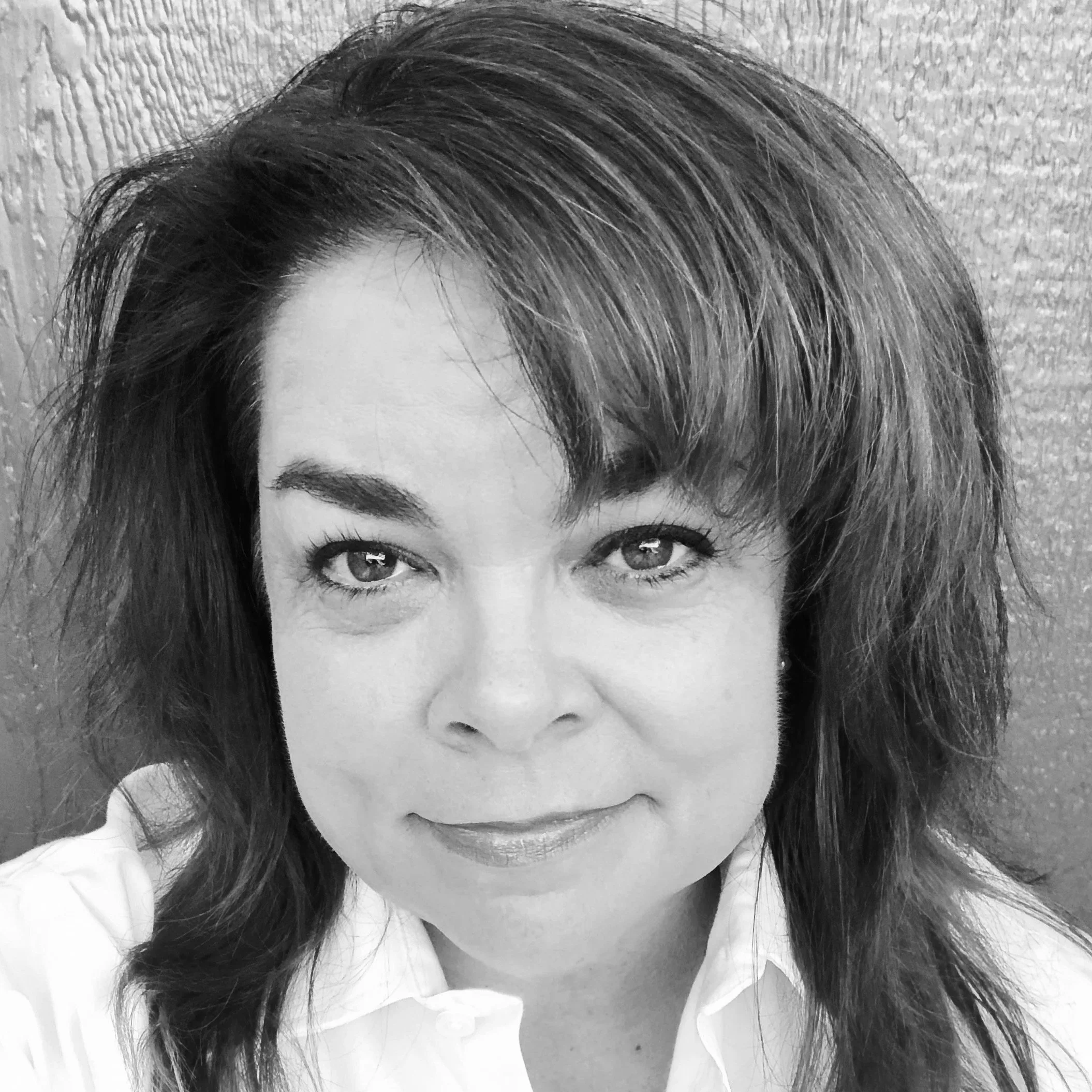 Black and white close-up portrait of a woman with shoulder-length wavy hair, smiling with visible teeth, wearing a collared shirt, standing against a textured wall background.