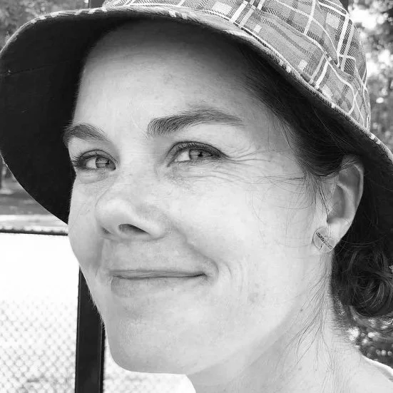 Black and white photo of a woman wearing a patterned hat, smiling softly, with a background that includes a fence and trees.