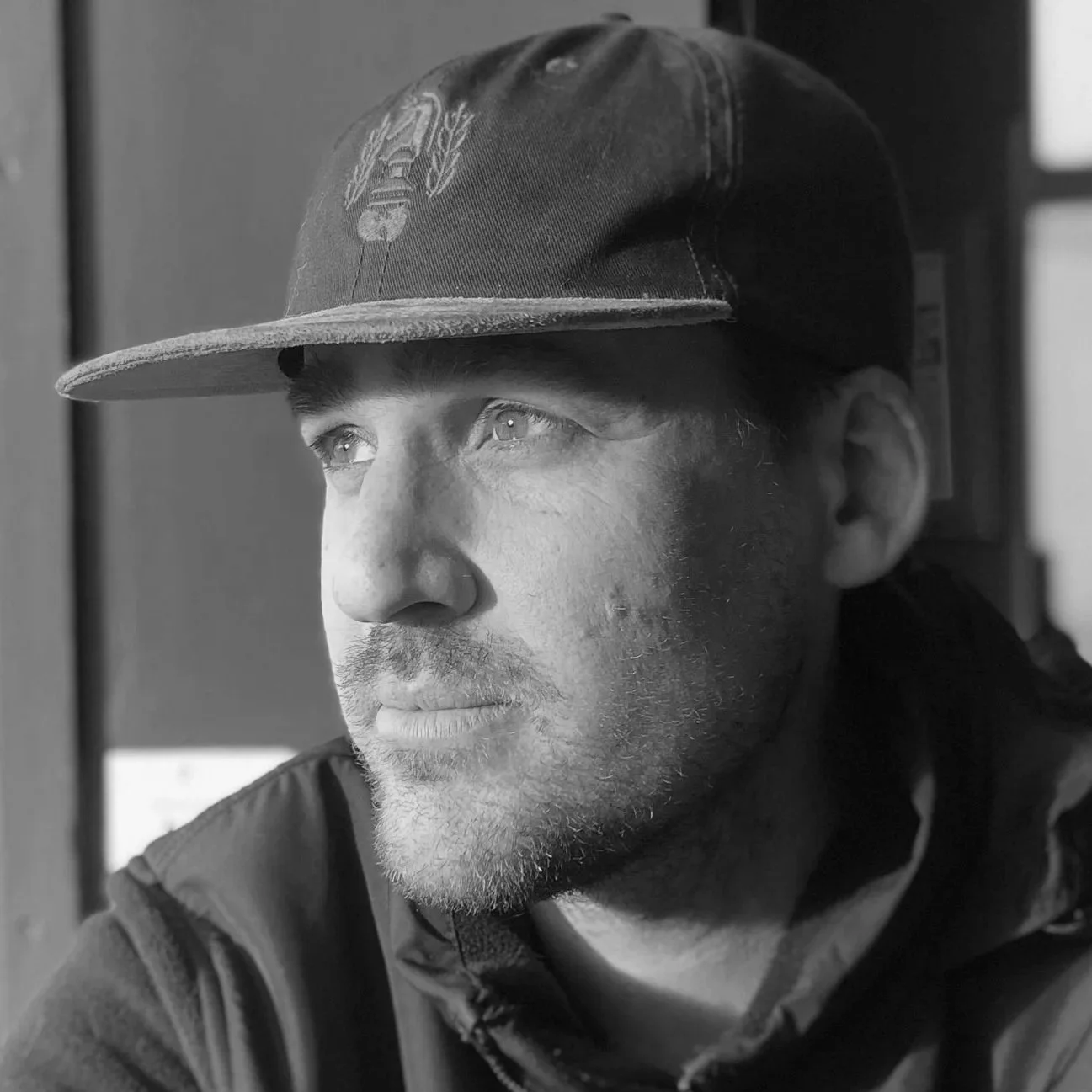 Close-up black and white photo of a man wearing a baseball cap, looking thoughtfully to the side.