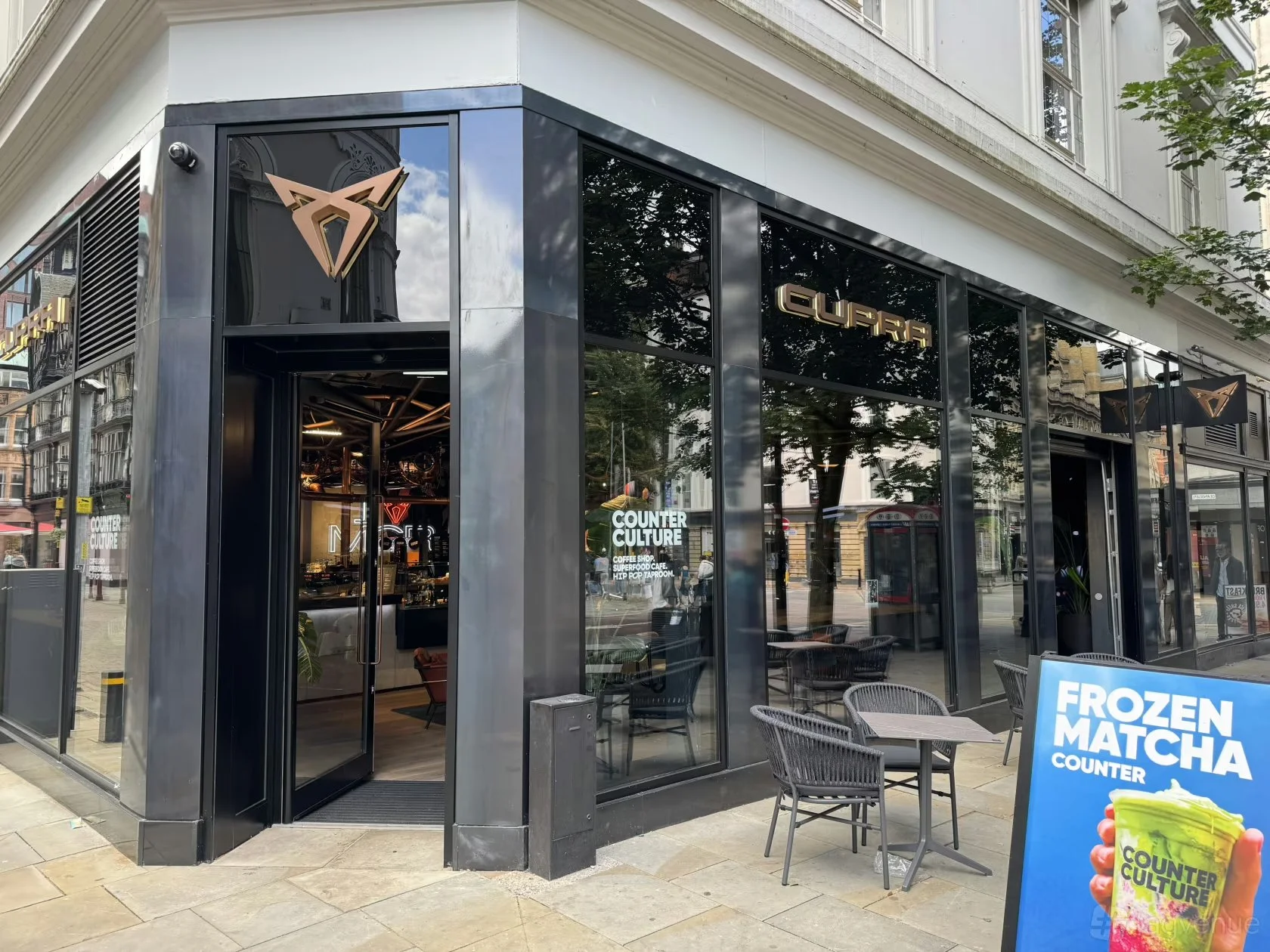 Storefront with illuminated sign reading 'CUPRA', displaying mannequins and cars inside, on a city street with small bollards and a sidewalk.