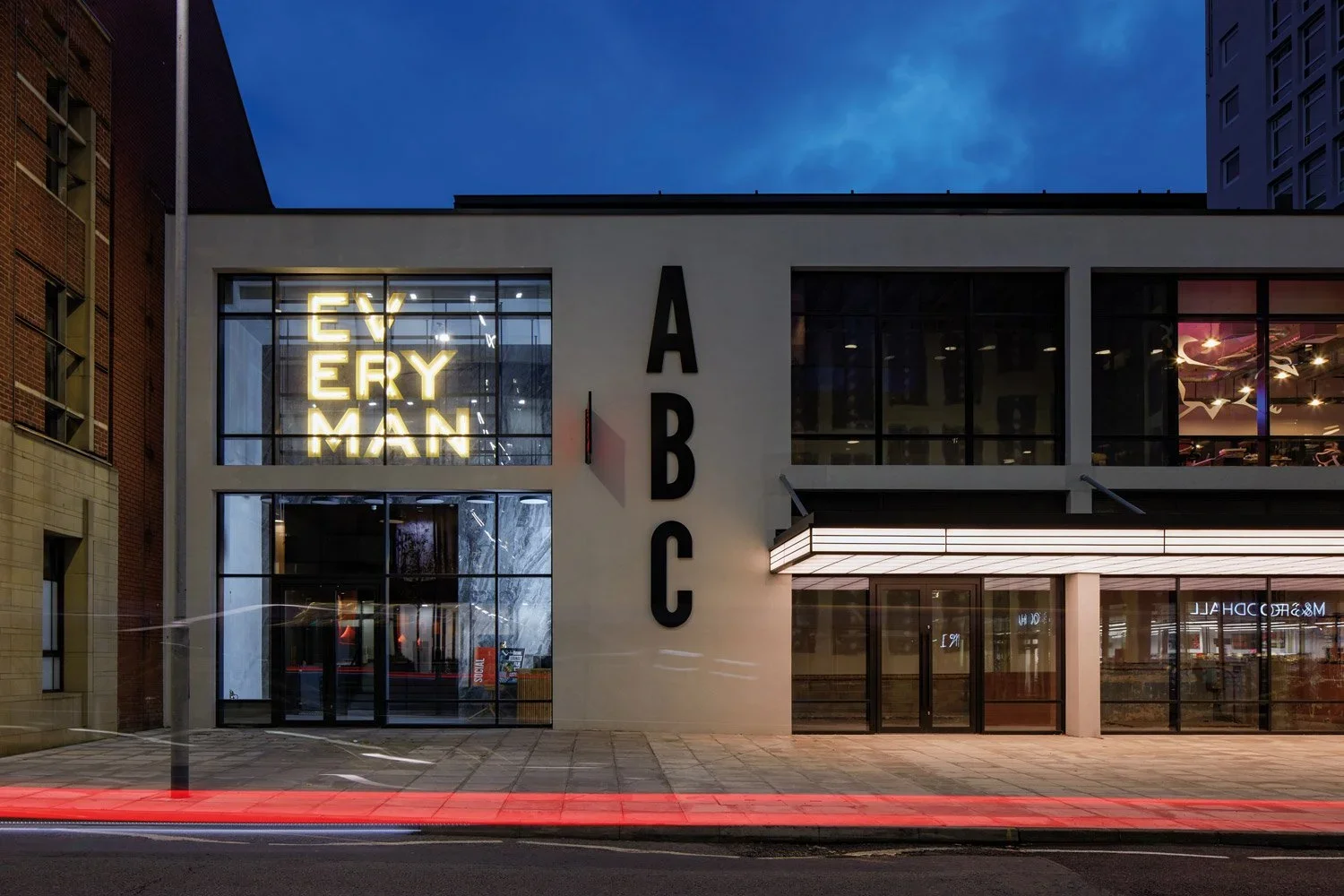 Modern building at night with large windows, a vertical sign spelling A B C, and illuminated yellow sign in the window that reads EY ERY MAN.