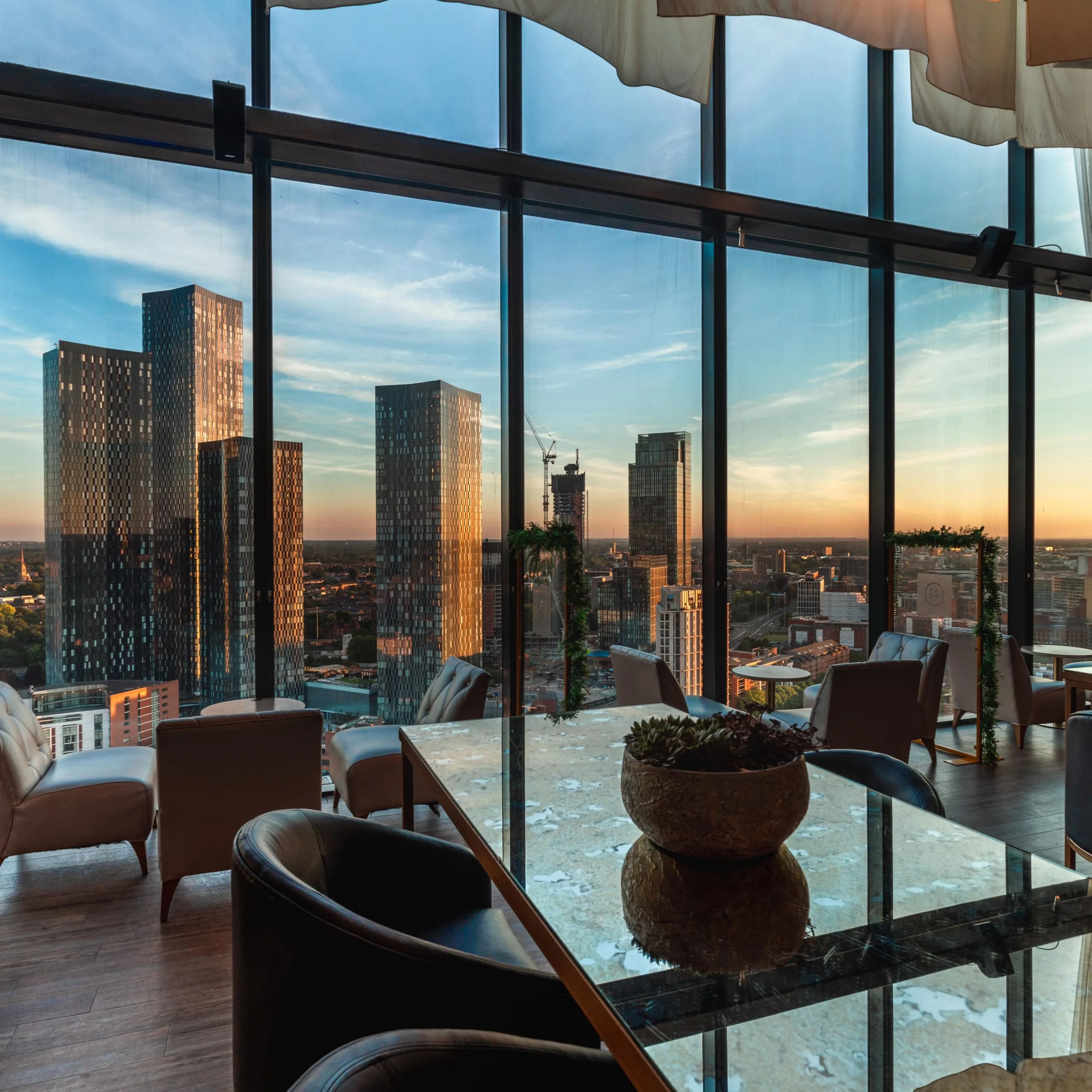 View of a city skyline through large glass windows, with high-rise buildings and a construction crane visible at sunset.