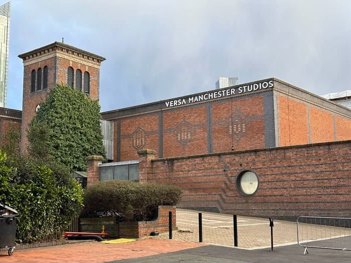 Exterior view of the Versa Manchester Studios building with a brick facade, surrounded by trees and a parking lot, with tall modern glass skyscrapers in the background on a sunny day.