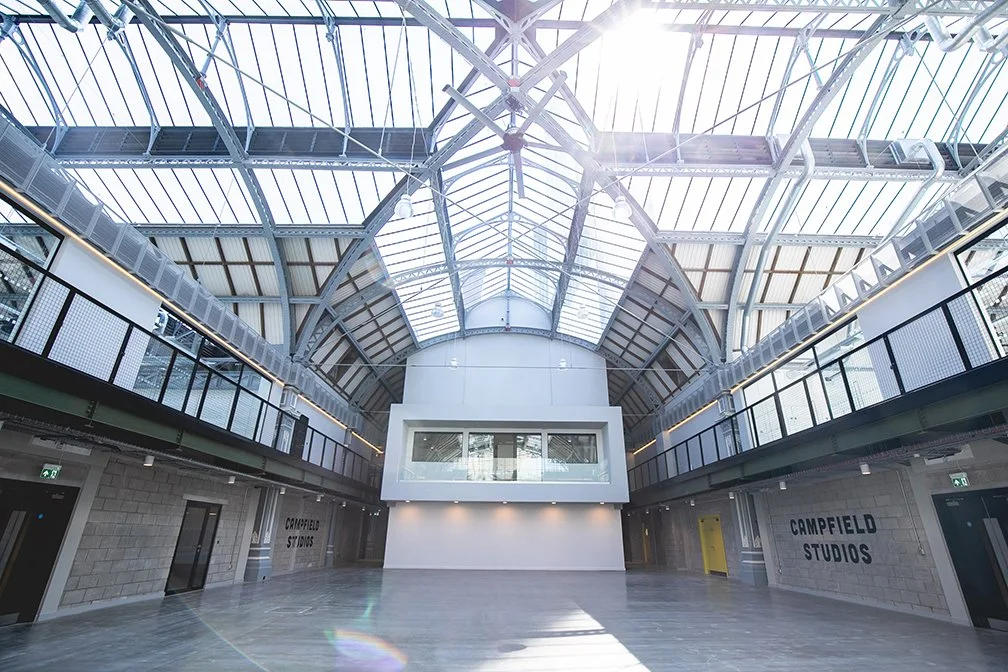 Interior of a modern, spacious building with a high glass ceiling, metal framework, and wooden accents, featuring a seating area with wooden pallet furniture and people walking around.