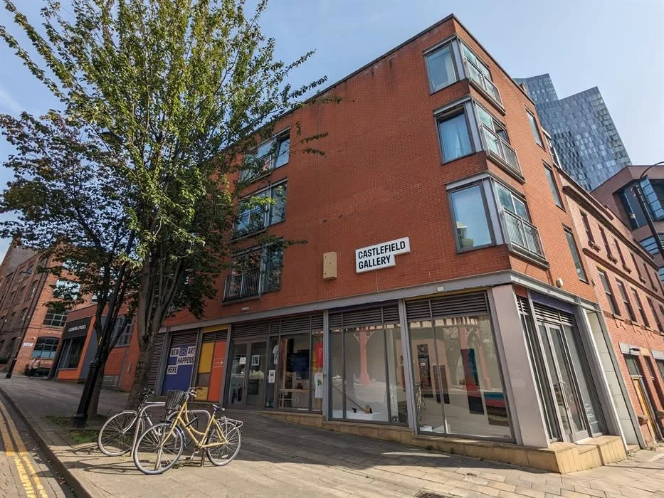 People sitting and standing outside a brick building with a sign for Castlefield Gallery Agency, some waiting at a bus stop or resting, a tree partially blocking the view, bicycles parked nearby, in daytime.