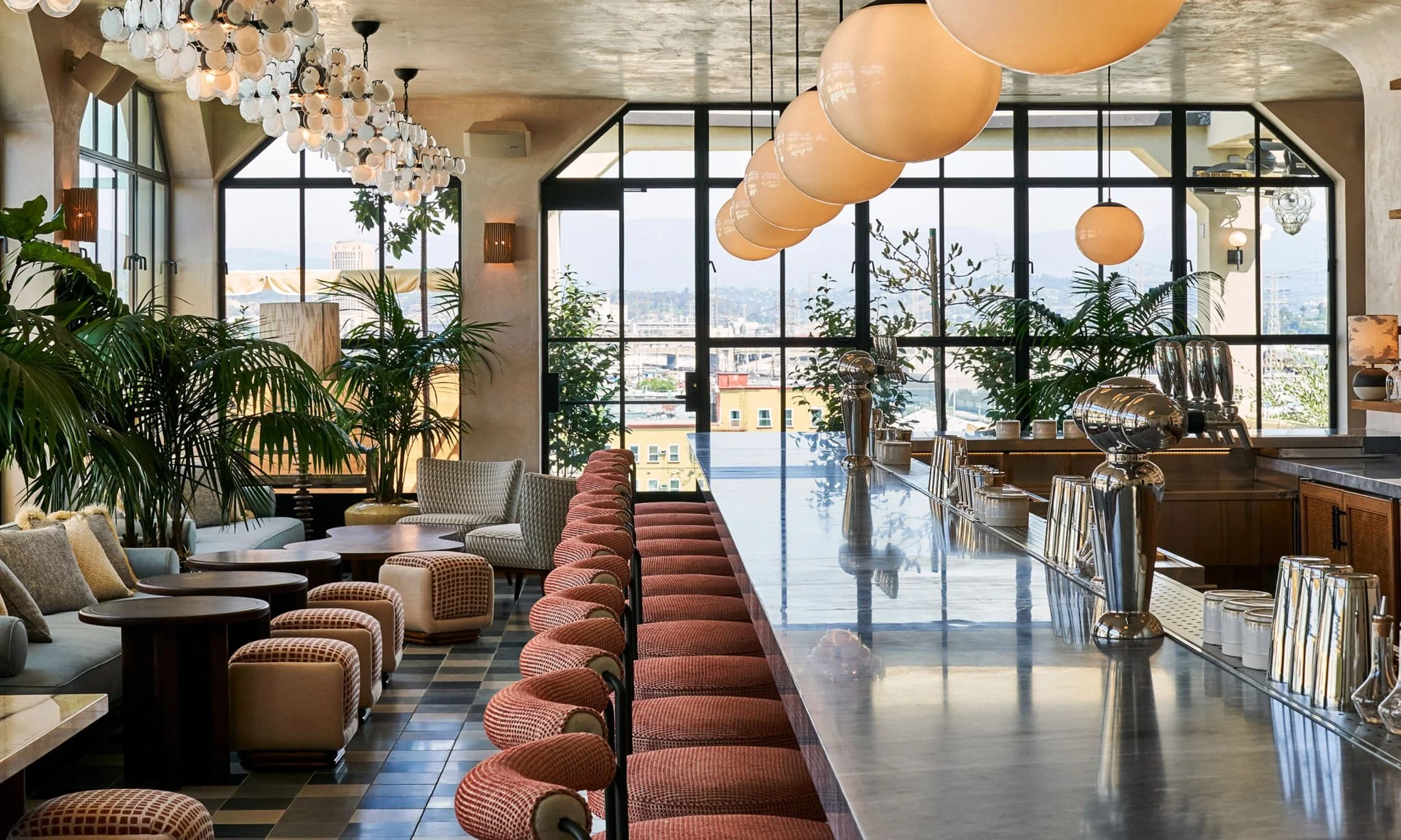 Modern restaurant interior with large windows, hanging spherical lights, a long bar counter with cocktail dispensers, and indoor plants.