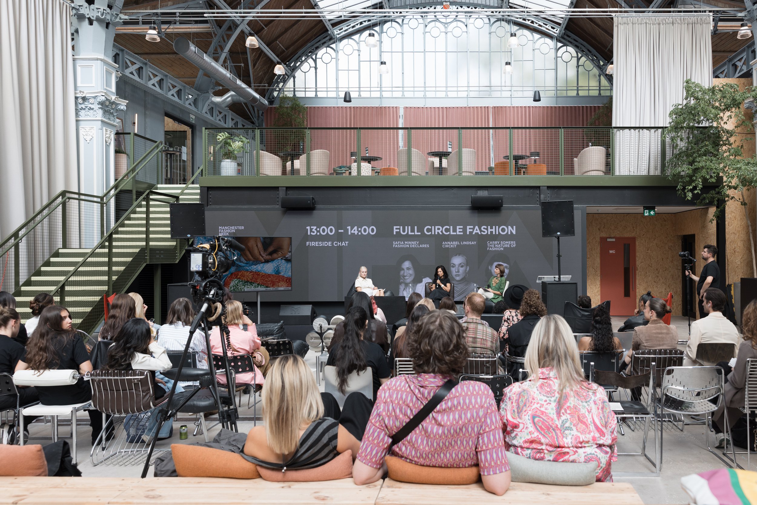 Audience attending a panel discussion at Manchester Fashion Week in a spacious industrial-style venue with high arched glass ceilings and metal beams. Four women sit on stage in front of a large screen displaying event details, and a cameraman record