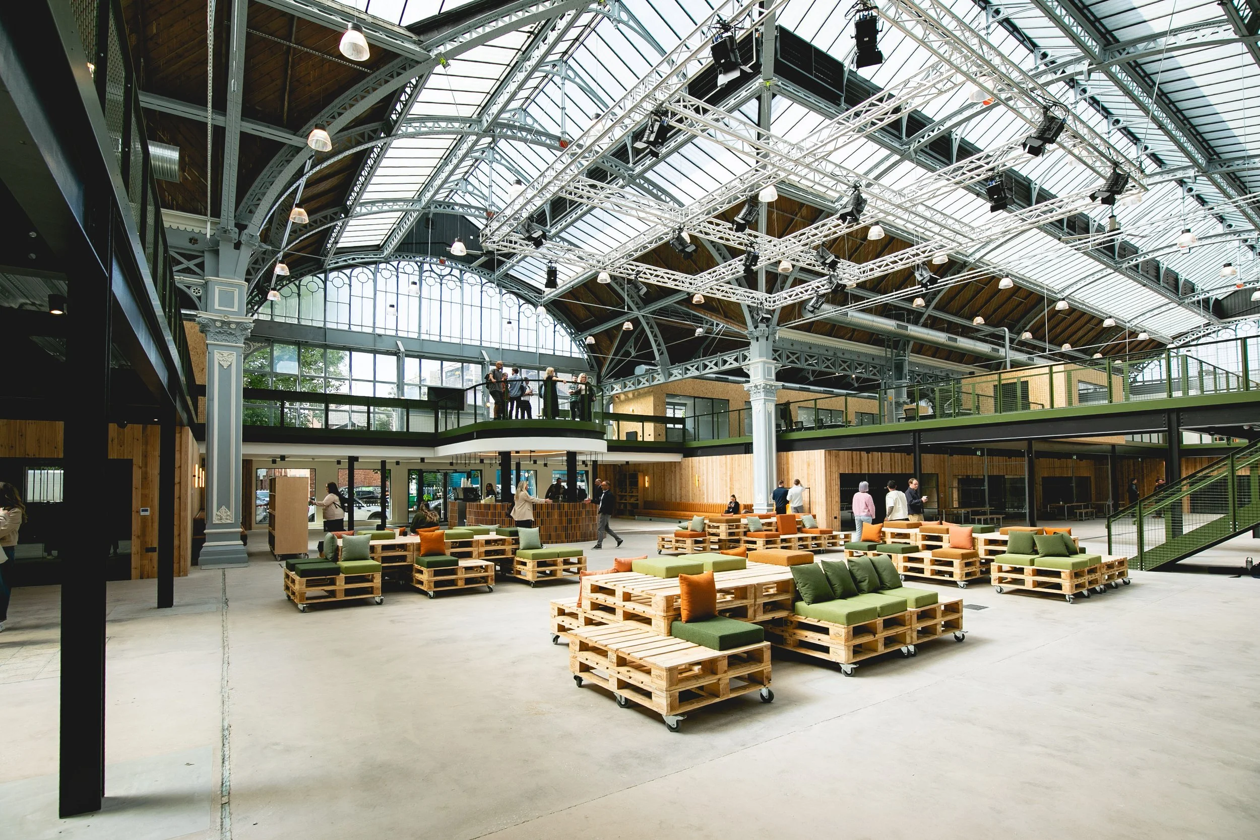 Interior of a modern, spacious building with a high glass ceiling, metal framework, and wooden accents, featuring a seating area with wooden pallet furniture and people walking around.