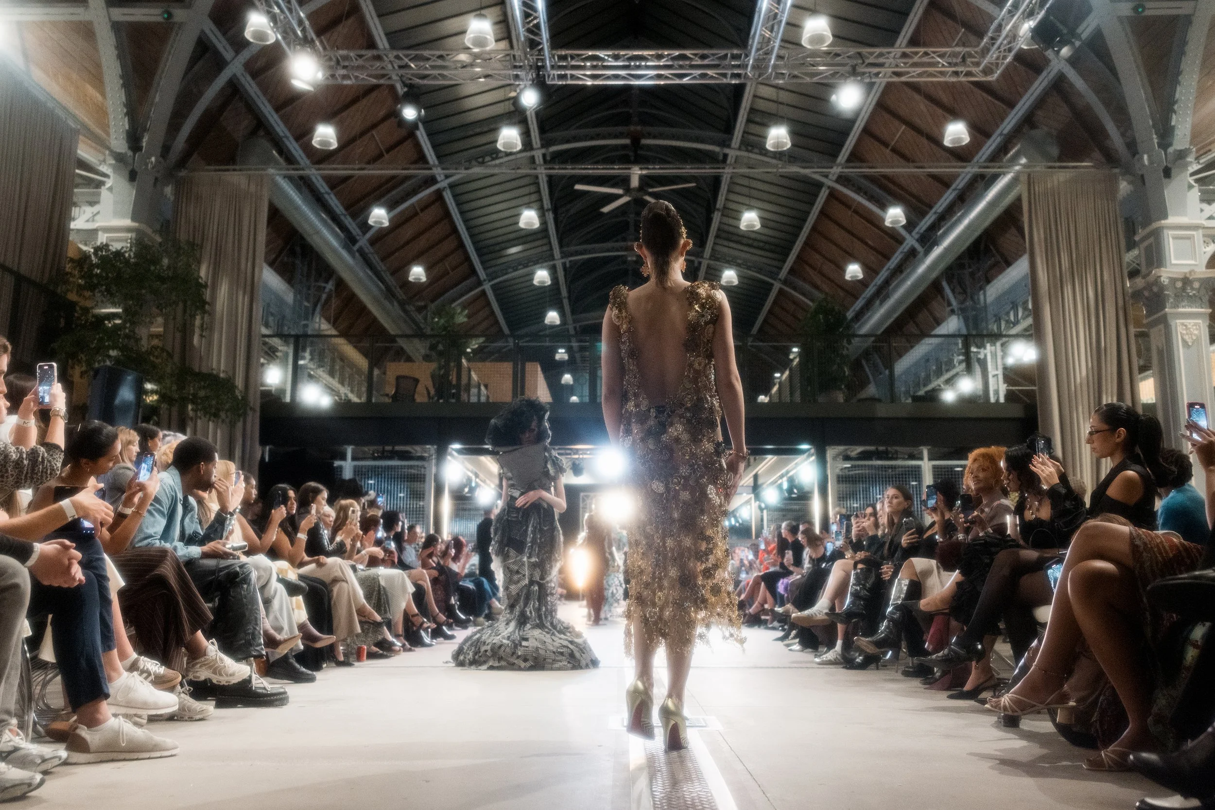 Fashion runway show in a spacious industrial-style venue with high ceilings and metal beams. A model in a sheer gown with gold embellishments walks down the runway, while the audience on both sides takes photos and watches.