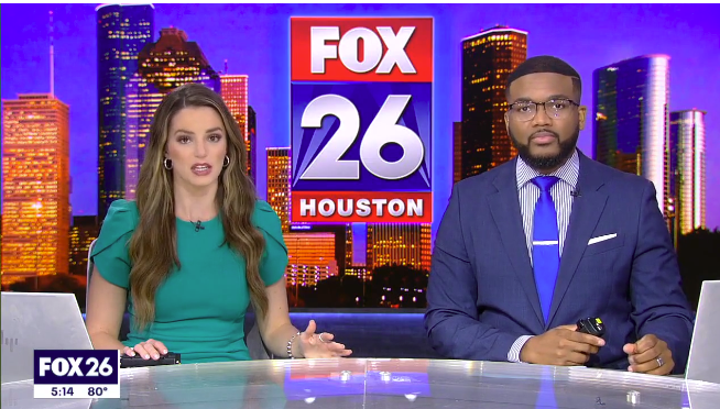Two news anchors sit at a desk in a television studio, with a digital background of the Houston city skyline and a large FOX 26 Houston logo displayed prominently behind them.