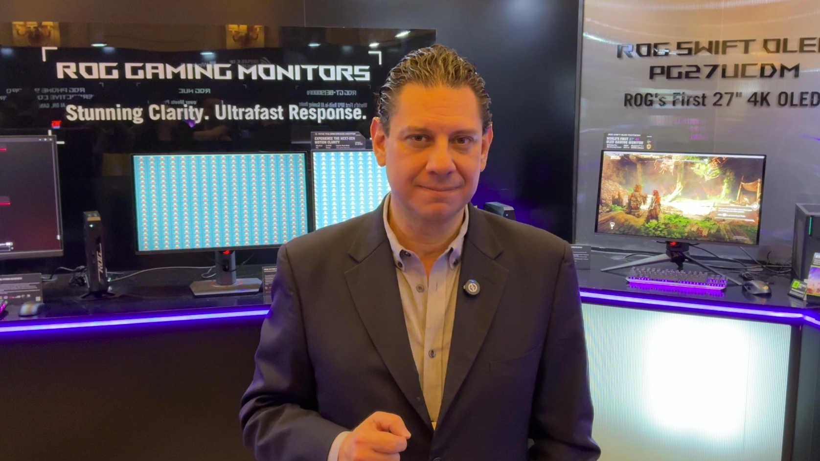 Juan Guevara Torres standing in front of a display booth with multiple computer monitors, advertising ROG gaming monitors and a 27-inch 4K OLED monitor at CES an electronics expo.