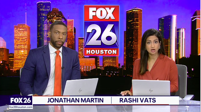 Two news anchors sitting at a desk with laptops, with the FOX 26 Houston logo and a city skyline background behind them. The male anchor on the left is wearing a navy pinstripe suit with an orange tie, and the female anchor on the right is wearing a reddish-brown blouse.