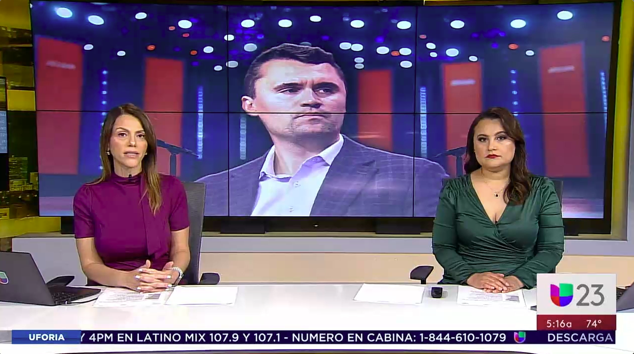 Two women news anchors sitting at a news desk with a large screen behind them showing a man's face. The screen has a red and blue background with lights and vertical lines. The U-23 logo and a weather report are visible on the lower right corner.