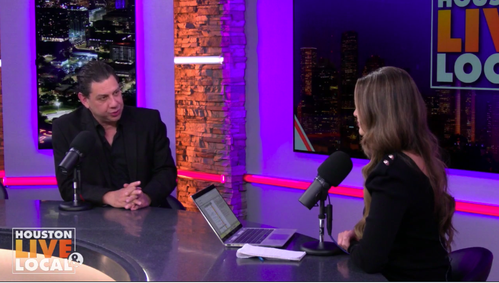 A radio studio with two people engaged in an interview. The man on the left has dark hair and is wearing a black suit. The woman on the right has long, wavy hair and is wearing a black top. They are seated at a table with microphones, a laptop, and a notebook. Behind them, a TV screen displays the show name "Houston Live Local" and a cityscape at night.