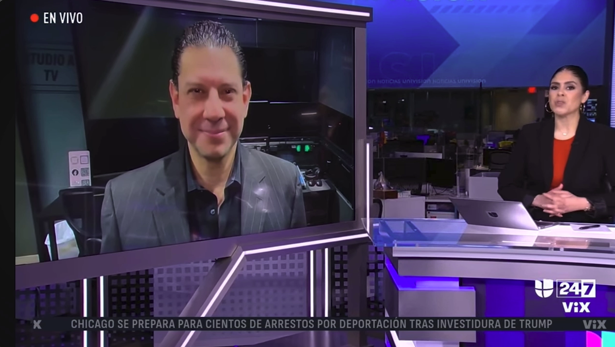 VIX Univision News broadcast studio with a female news anchor sitting at a desk with a laptop, and Juan Guevara Torres appearing on a large screen via video call, wearing a gray pinstripe suit.