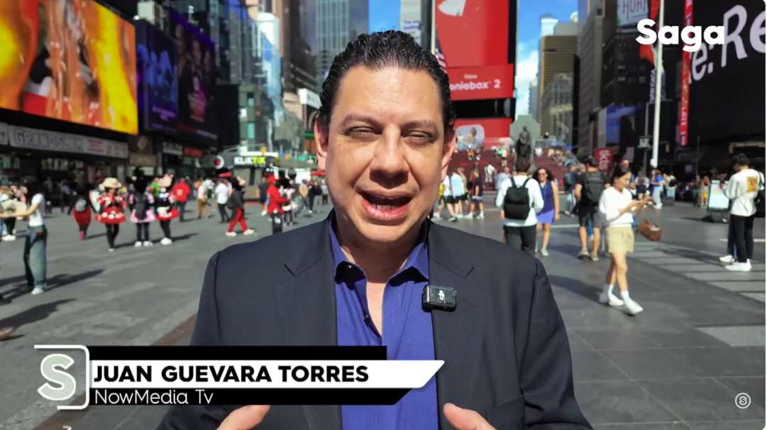 Man in a suit speaking in Times Square, New York City, with crowds, electronic billboards, and buildings in the background.