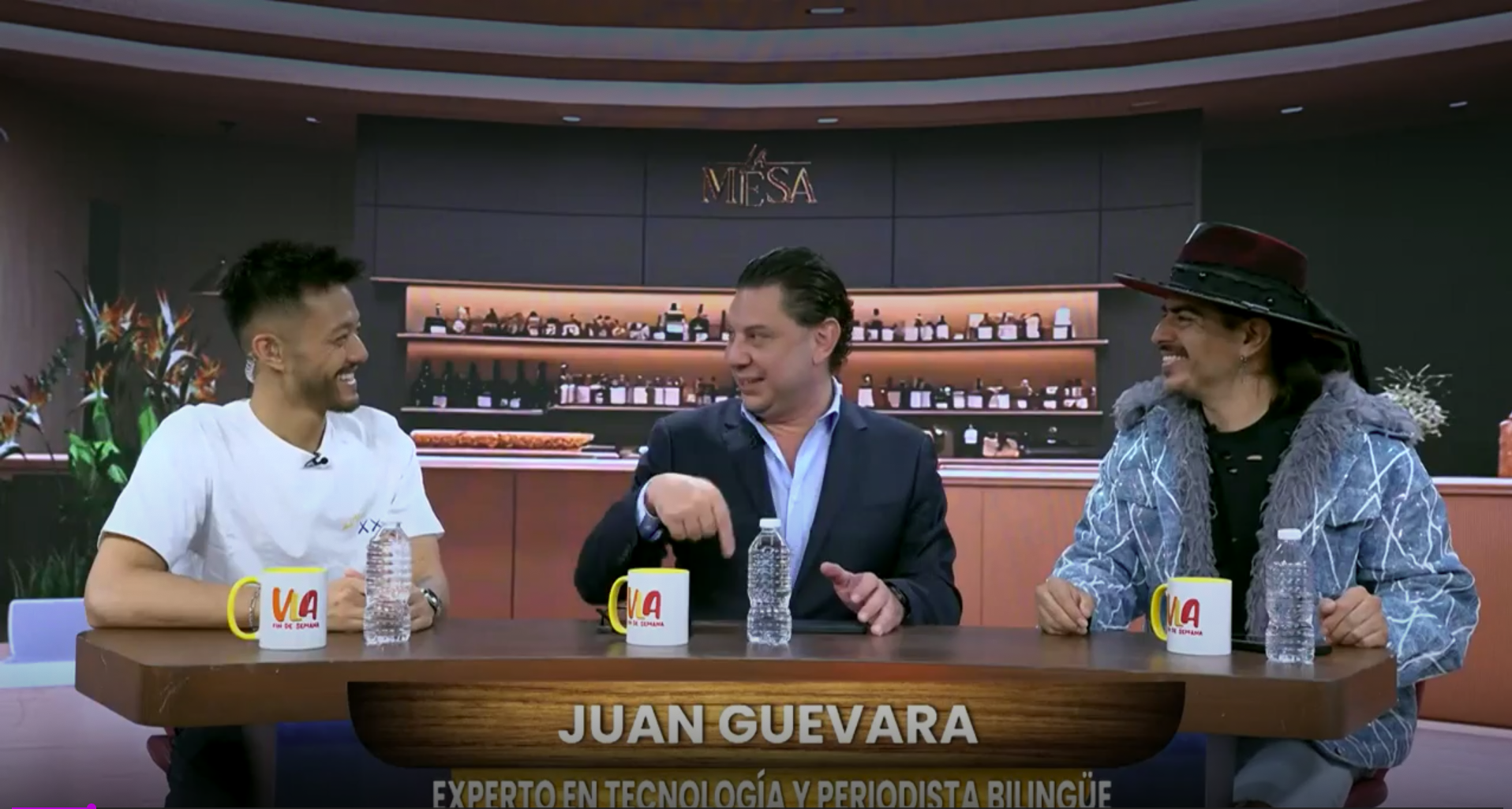 Three men sitting at a table in a TV studio with a bar background, smiling and talking. The man in the middle points to himself. The table has water bottles and yellow coffee mugs. Text on screen reads "JUAN GUEVARA, EXPERTO EN TECNOLOGÍA Y PERIODISTA BILINGÜE."