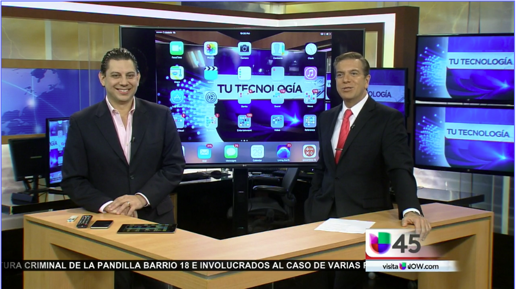 Juan Guevara Tech expert and Raul Peimbert news anchor in suits standing behind a wooden desk with multiple screens displaying technology graphics, in a television studio setting.