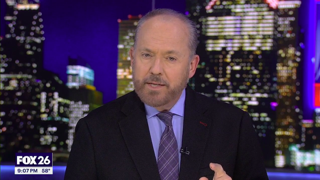 Tom Zizka news anchor in a black suit, purple shirt, and patterned tie, with city skyline backdrop at night, on FOX 26.