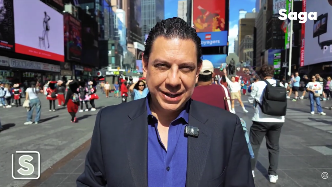 Man in blue shirt and dark blazer standing in busy Times Square with crowds and digital billboards in New York City.