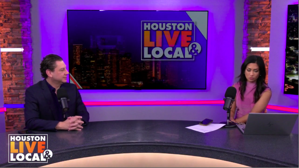Two people sit at a table in a radio studio with Coca-Cola themed background, microphones, and a large screen displaying the 'Houston Live Local' logo.