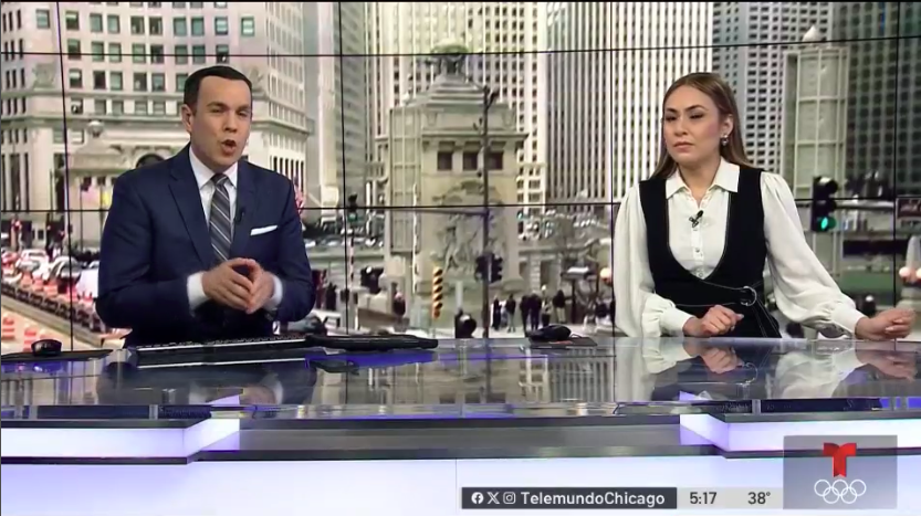 Two news anchors sit at a desk in a TV studio, with a large window behind them showing a cityscape with tall buildings and a statue. The male anchor is speaking while the female anchor listens. A TelemundoChicago logo and Olympic rings are visible at the bottom of the screen.