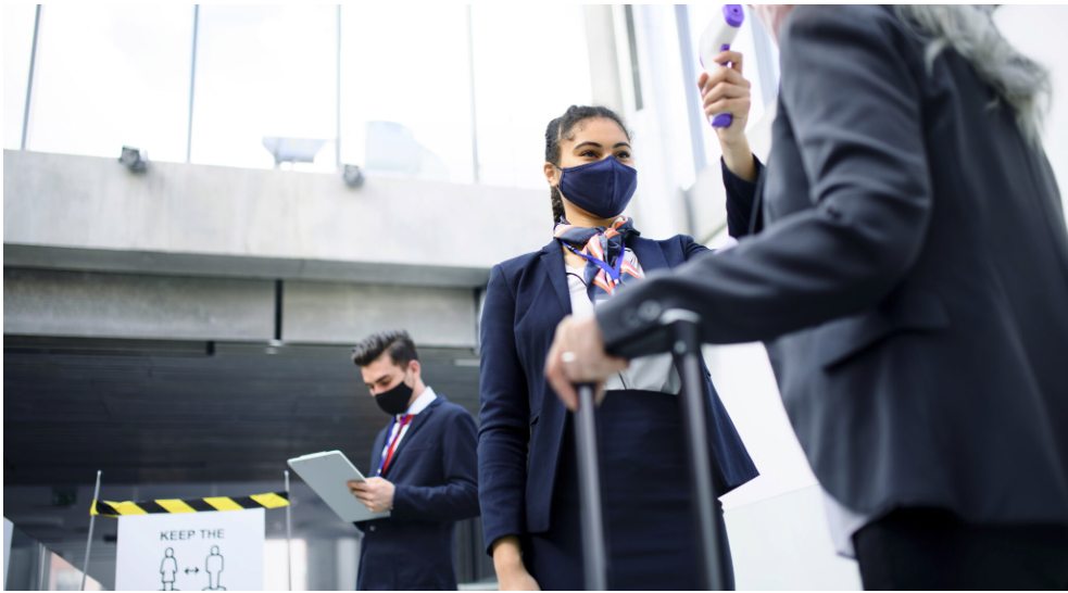 Two women wearing suits and face masks, one holding a travel handle, at an airport security checkpoint. A man in a suit and mask stands in the background, looking at a tablet device.