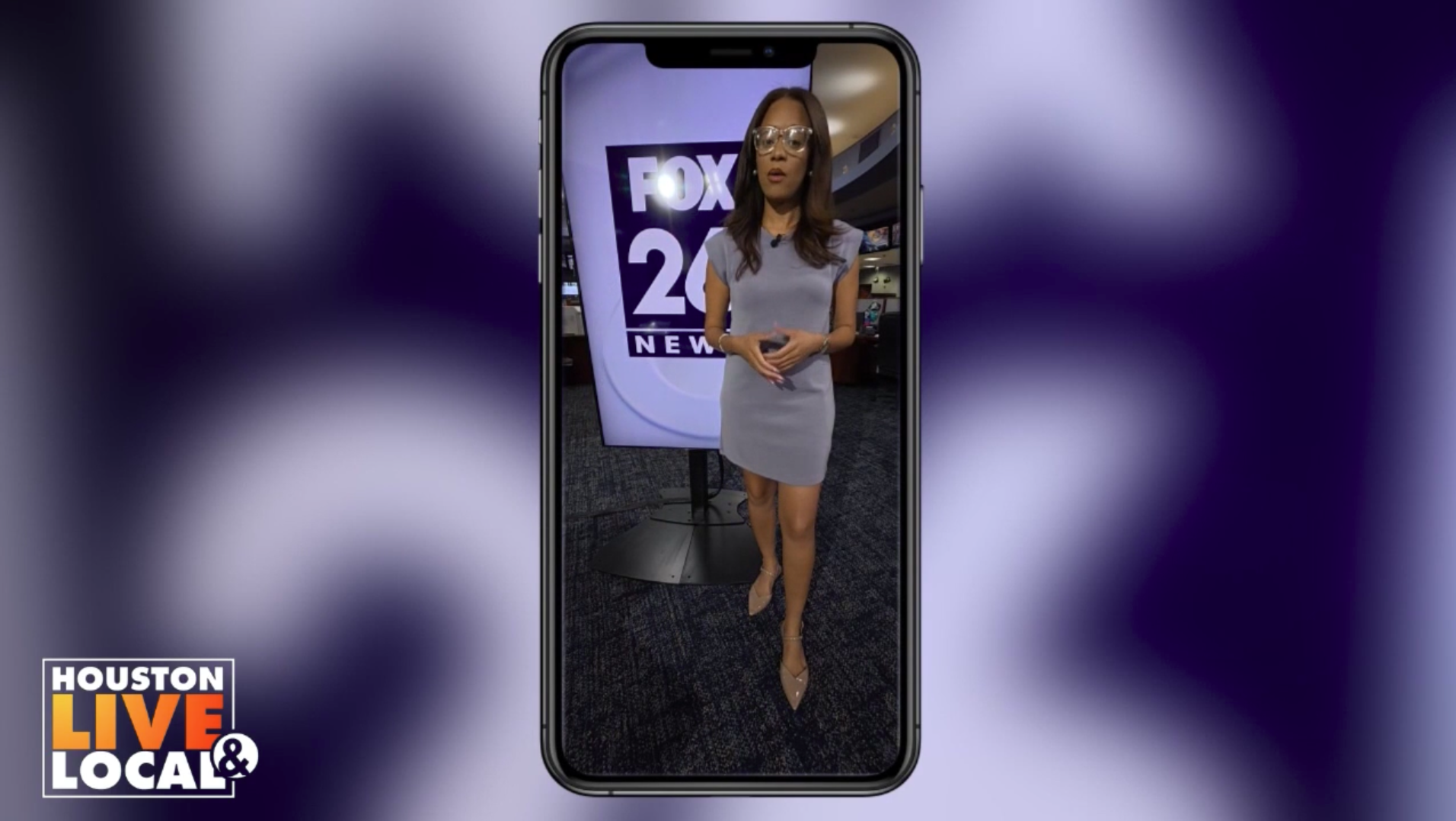 A woman in a grey dress and heels appearing on a large smartphone screen with a FOX 26 News background, in a newsroom setting.