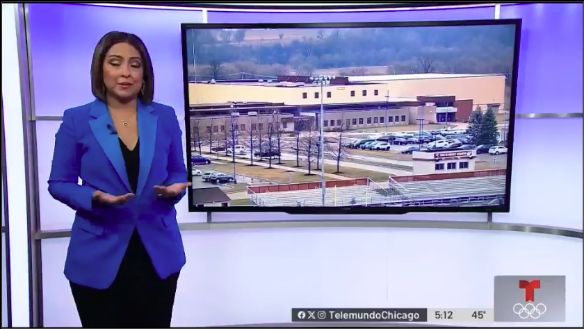 Television news anchor in a blue blazer presenting in front of a large screen showing an aerial view of a school or industrial building with parking lots and trees.
