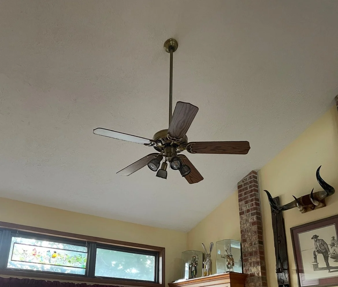 Ceiling fan with five wooden blades and four light fixtures in a room with beige walls, brick pillar, and framed artwork.