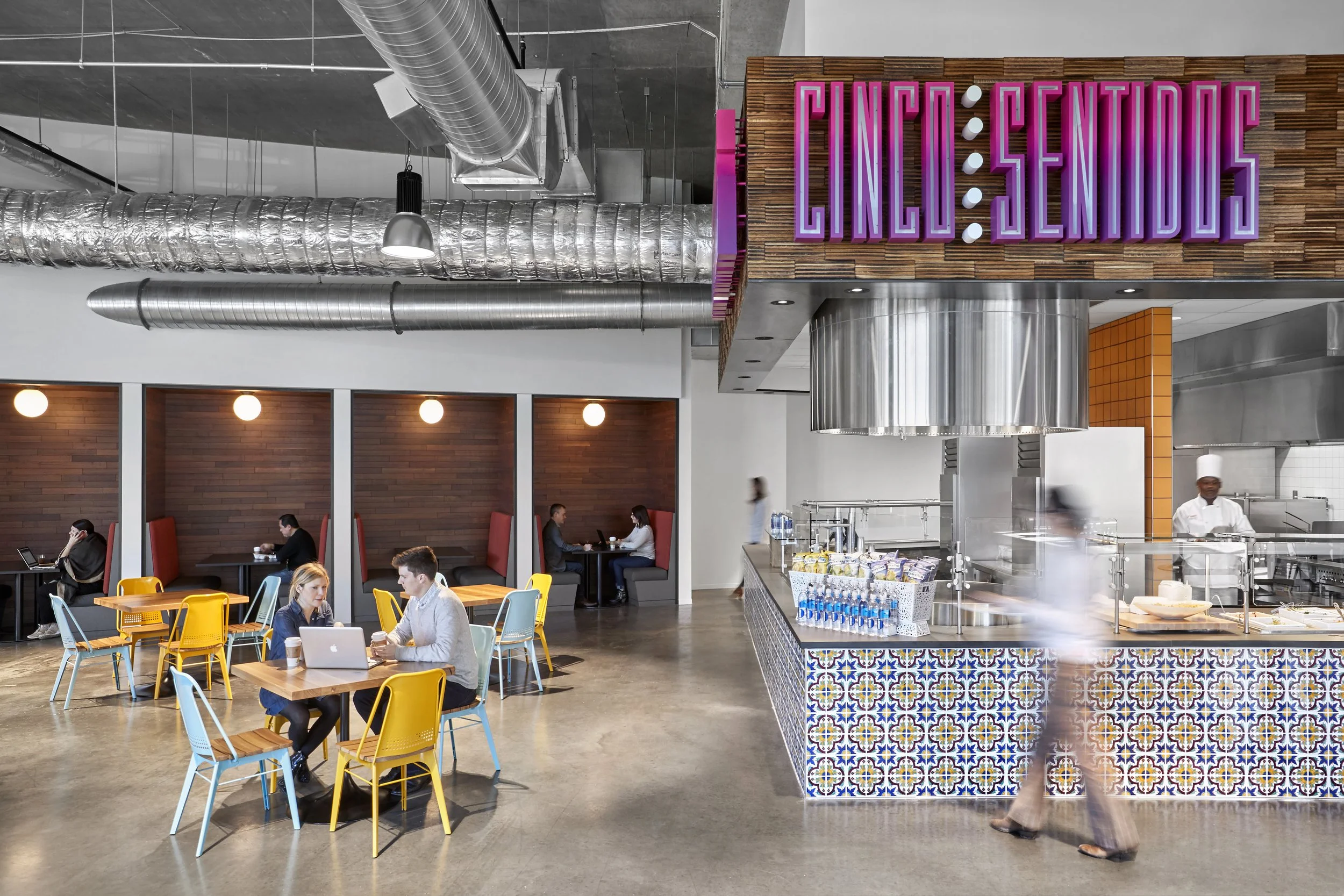 Interior of a modern cafeteria with colorful chairs, some people working on laptops or having conversations, a kitchen area with a chef, and a neon sign that reads 'Cinco Sentidos' in purple and pink