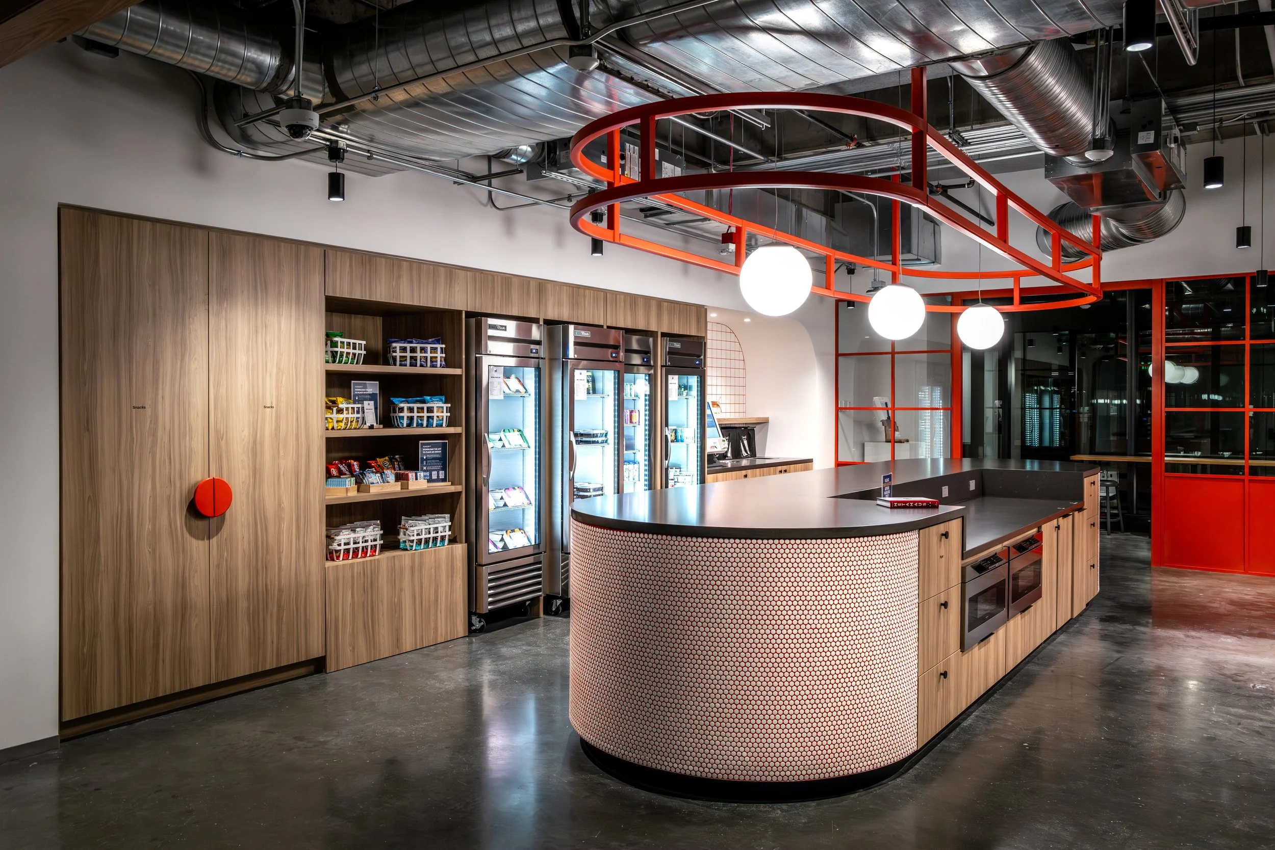 Modern coffee shop counter with wooden cabinets, refrigerated display case, vending machines, and red ceiling fixtures with three white globe lights, in an industrial-style space.