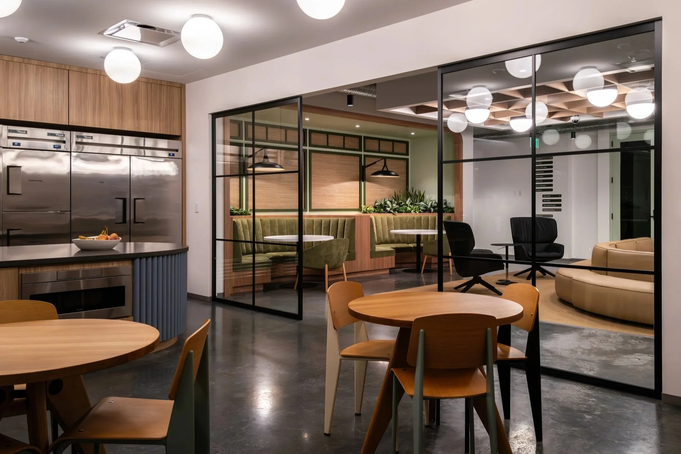 Modern interior cafe with wooden tables, green upholstered booths, black and beige chairs, and a kitchenette area with stainless steel appliances and a bowl of fruit.