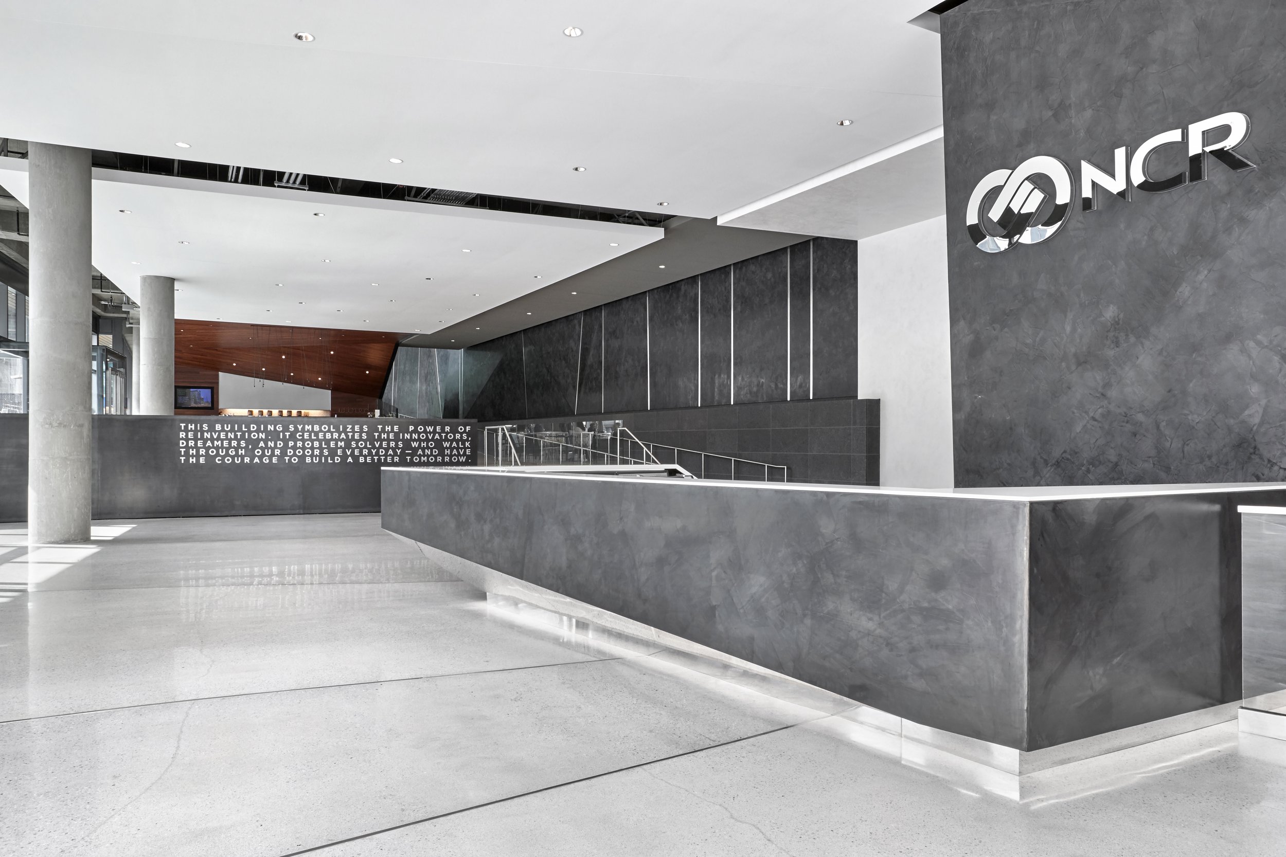 Modern lobby interior with gray and black walls, a curved staircase with metal railings, and a logo that reads 'NCR' on the wall.