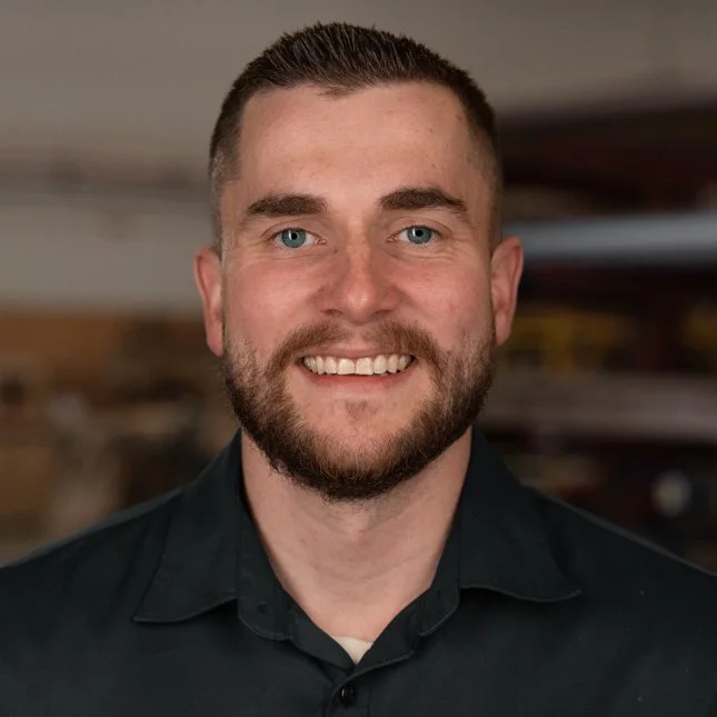 Portrait of a smiling man with short brown hair, a beard, and blue eyes, wearing a black collared shirt with a blurred indoor background.