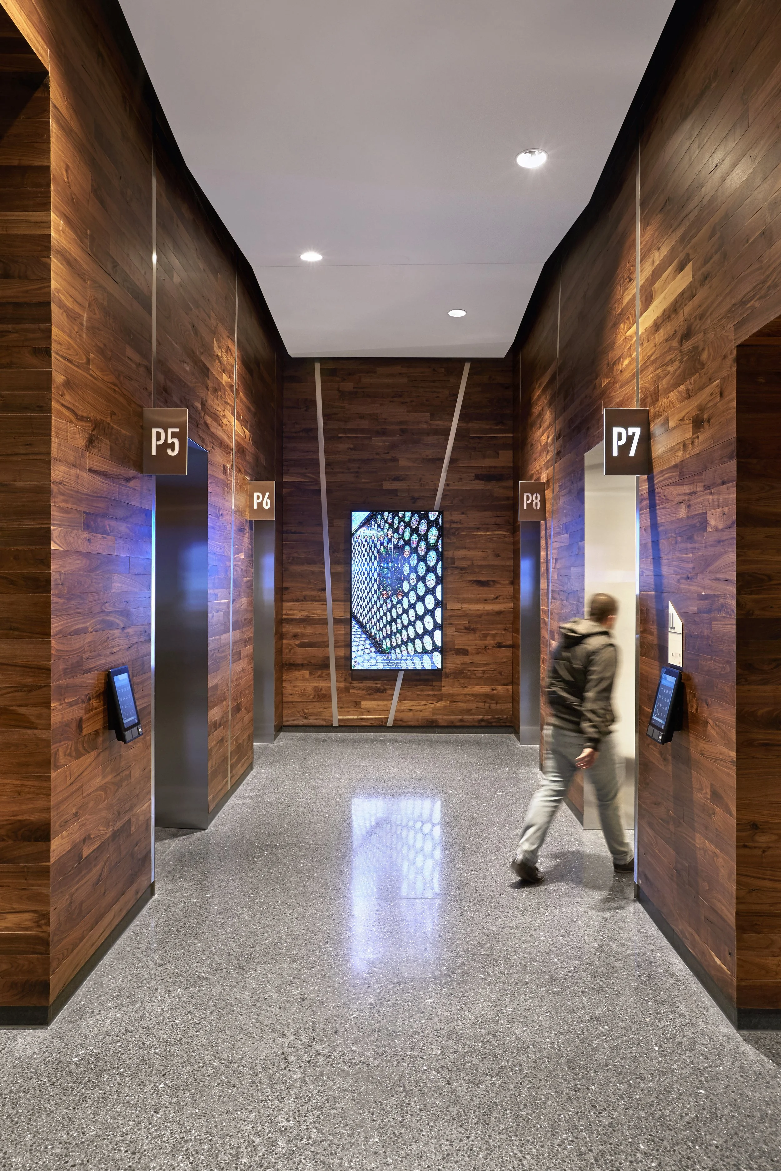 An indoor hallway with wooden walls and a gray speckled floor, featuring four open elevator doors labeled P5, P6, P8, and P7, with digital panels beside each, and a man walking near the P7 door.