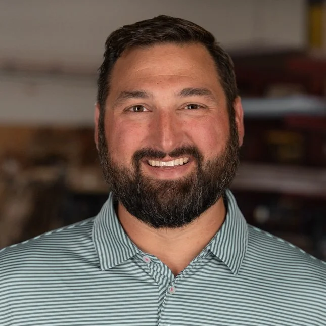 A bearded man with short dark hair wearing a light blue and white striped button-up shirt, smiling at the camera in an indoor setting.
