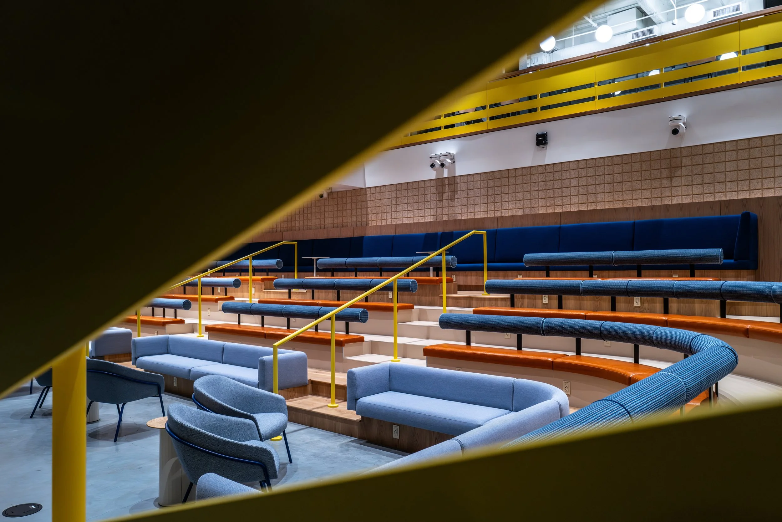 Interior of a modern auditorium or lecture hall with tiered blue seating, some seating areas with orange upholstery, and a few small tables. The photo is taken through a yellow structure framing part of the view.