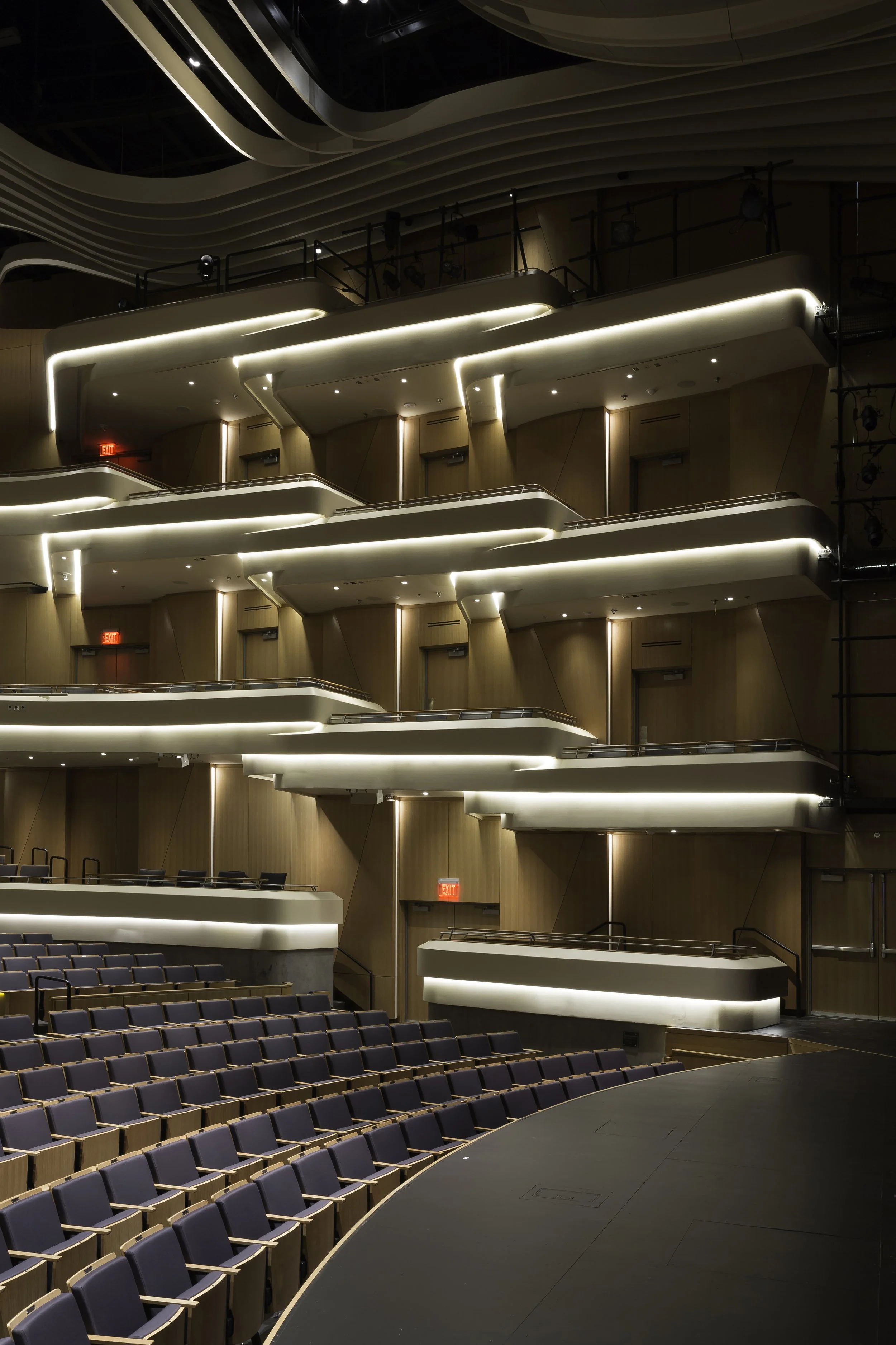 Interior of a modern theater or concert hall with multiple balcony levels, illuminated with soft lighting, wood paneling on walls, and many empty seats in the auditorium.