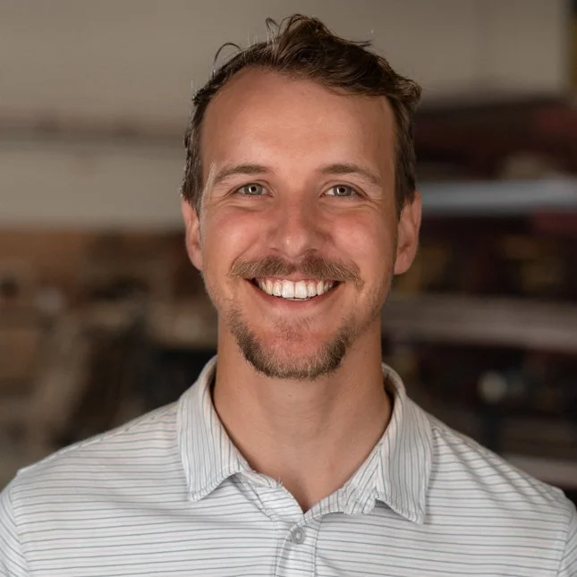 A smiling man with short brown hair, a beard, and mustache, wearing a light-colored striped shirt, in an indoor setting.