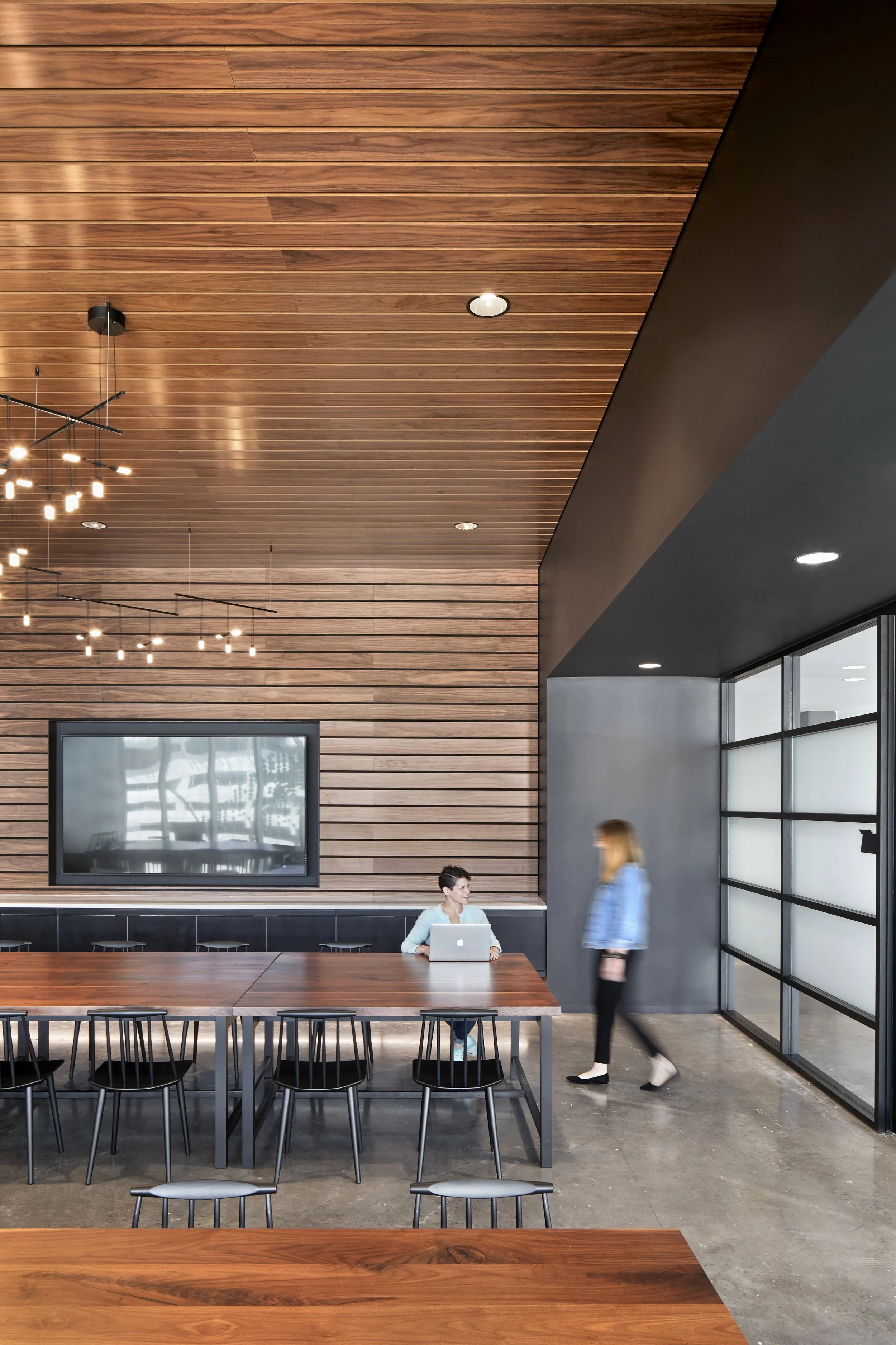 Interior of a modern dining or conference area with wooden ceiling and walls, a large flat-screen TV, and a long wooden table surrounded by black chairs. A woman sits at the table with a laptop, while another woman walks by.