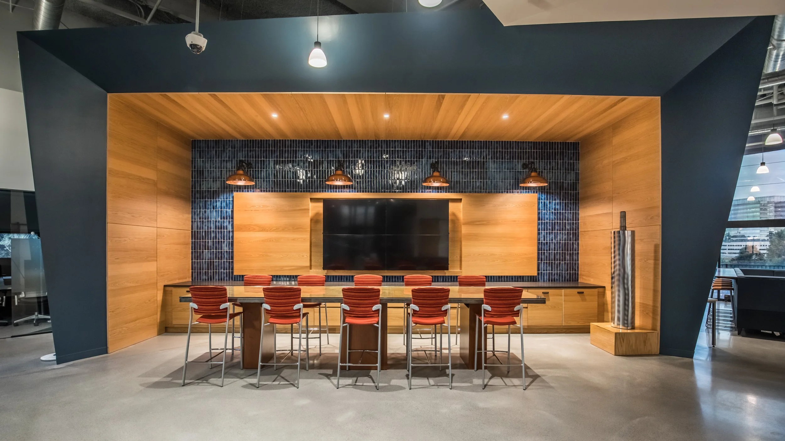 Modern conference room with wooden walls, a large flat-screen TV, red chairs around a wooden table, and a decorative column, in an office building.