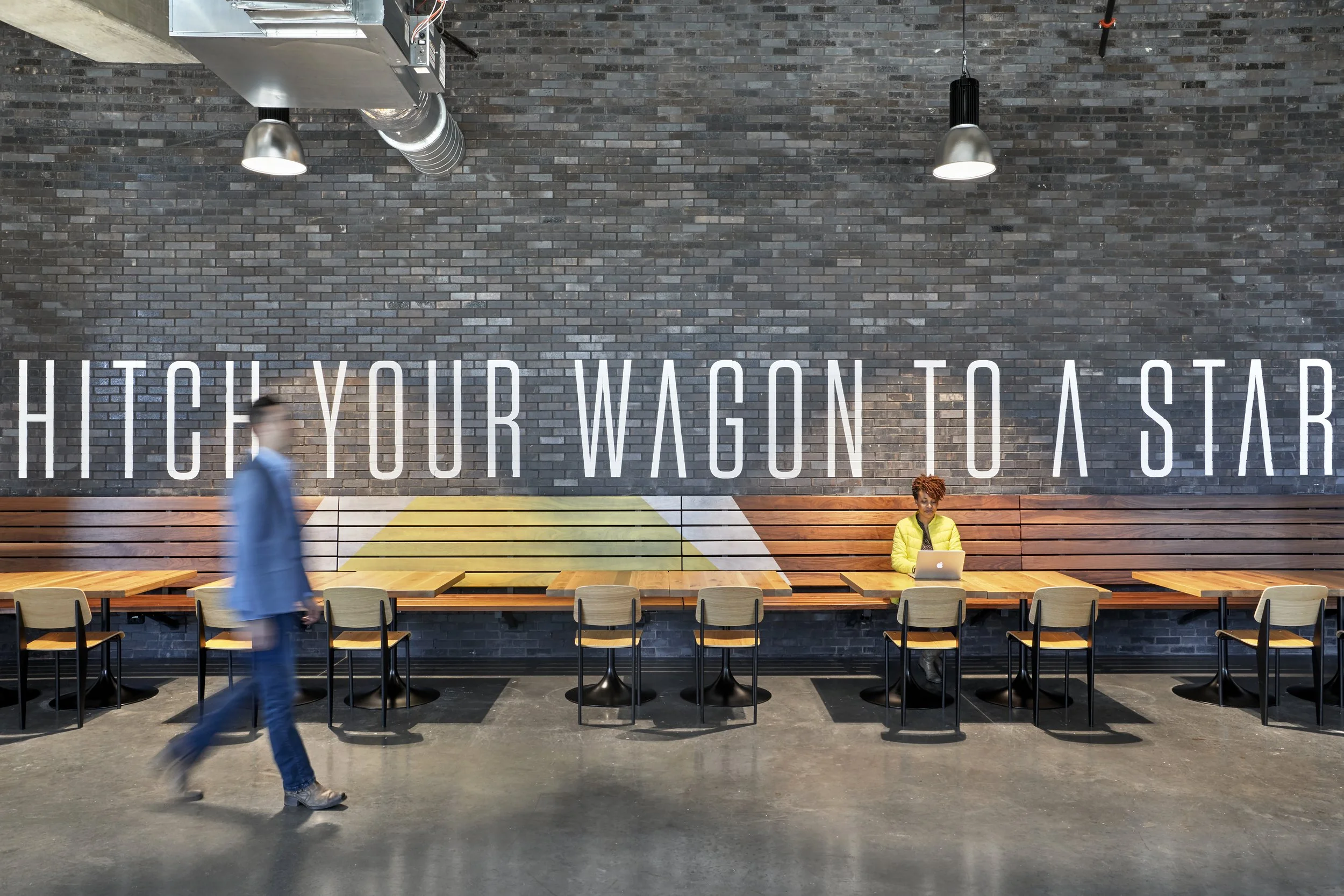 Interior of a modern cafe or communal space with a brick wall featuring large white letters that read "HITCH YOUR WAGON TO A STAR". There are wooden tables and chairs along the wall, one person sitting with a laptop and another person walking past, a