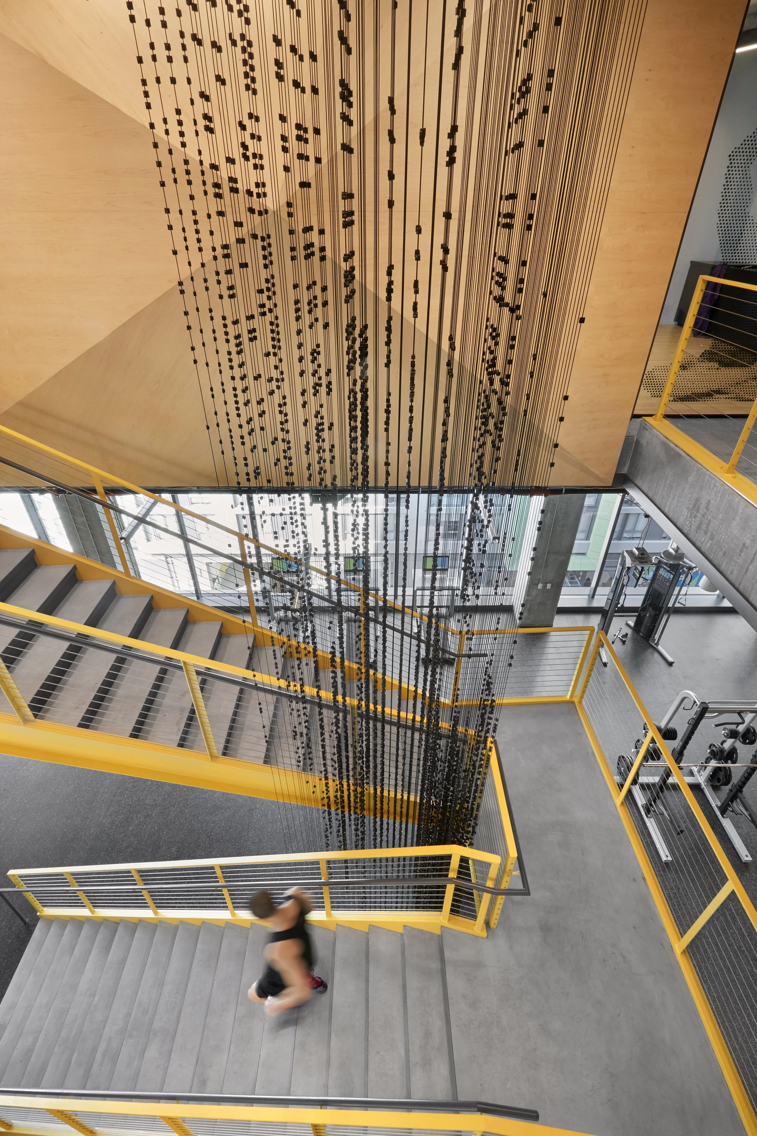 A person in athletic clothing walking down stairs in a modern building with yellow railings and a decorative hanging installation from the ceiling.